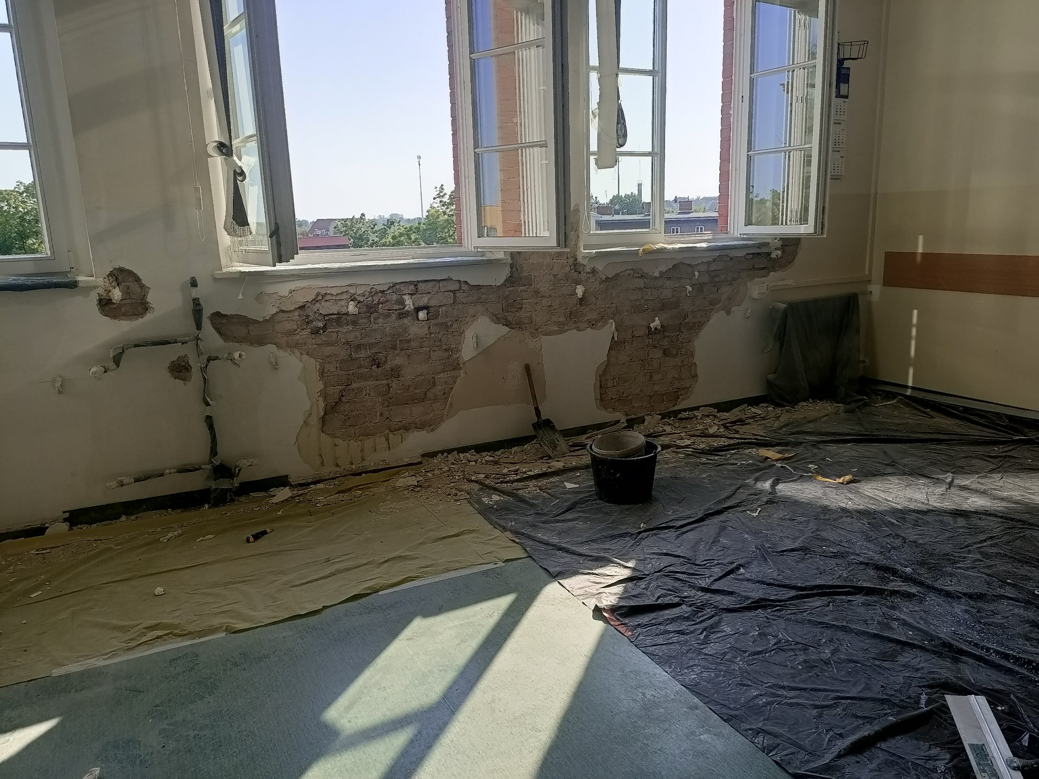 Pokój w trakcie skuwania tynku, odsłonięta cegła, zabezpieczona podłoga folią. Widoczne narzędzia i wiadro. Przygotowanie do dalszych prac remontowych.