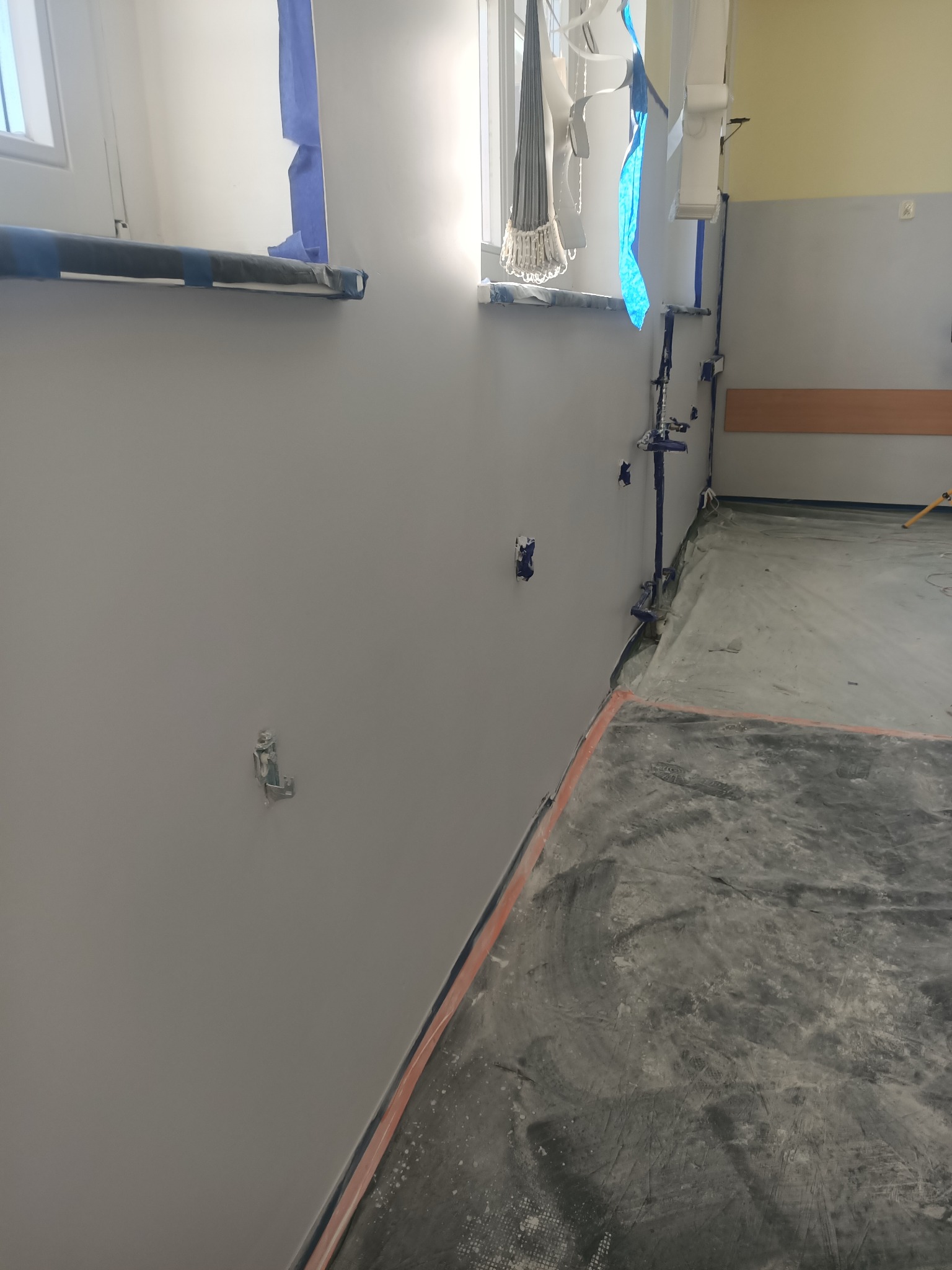 Ściana po szpachlowaniu i malowaniu, zabezpieczona folią i taśmą malarską. Widoczne puszki elektryczne i osłonięte okno. Przygotowanie do dalszych prac wykończeniowych.