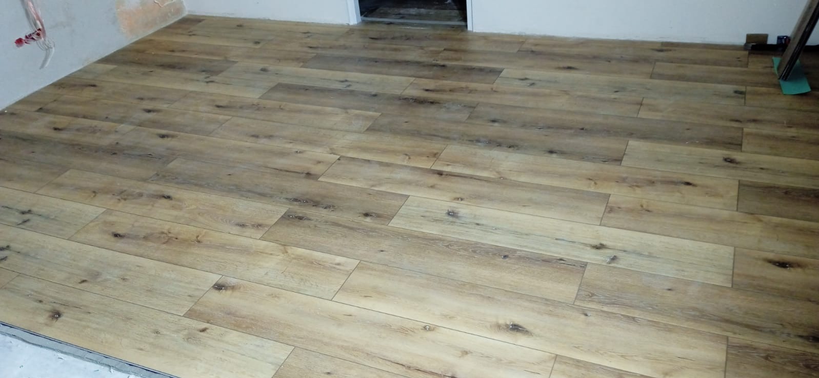 Ułożona podłoga z paneli imitujących drewno w jasnym odcieniu, widoczne niedokończone ściany i fragment progu. Układ paneli w jodełkę.