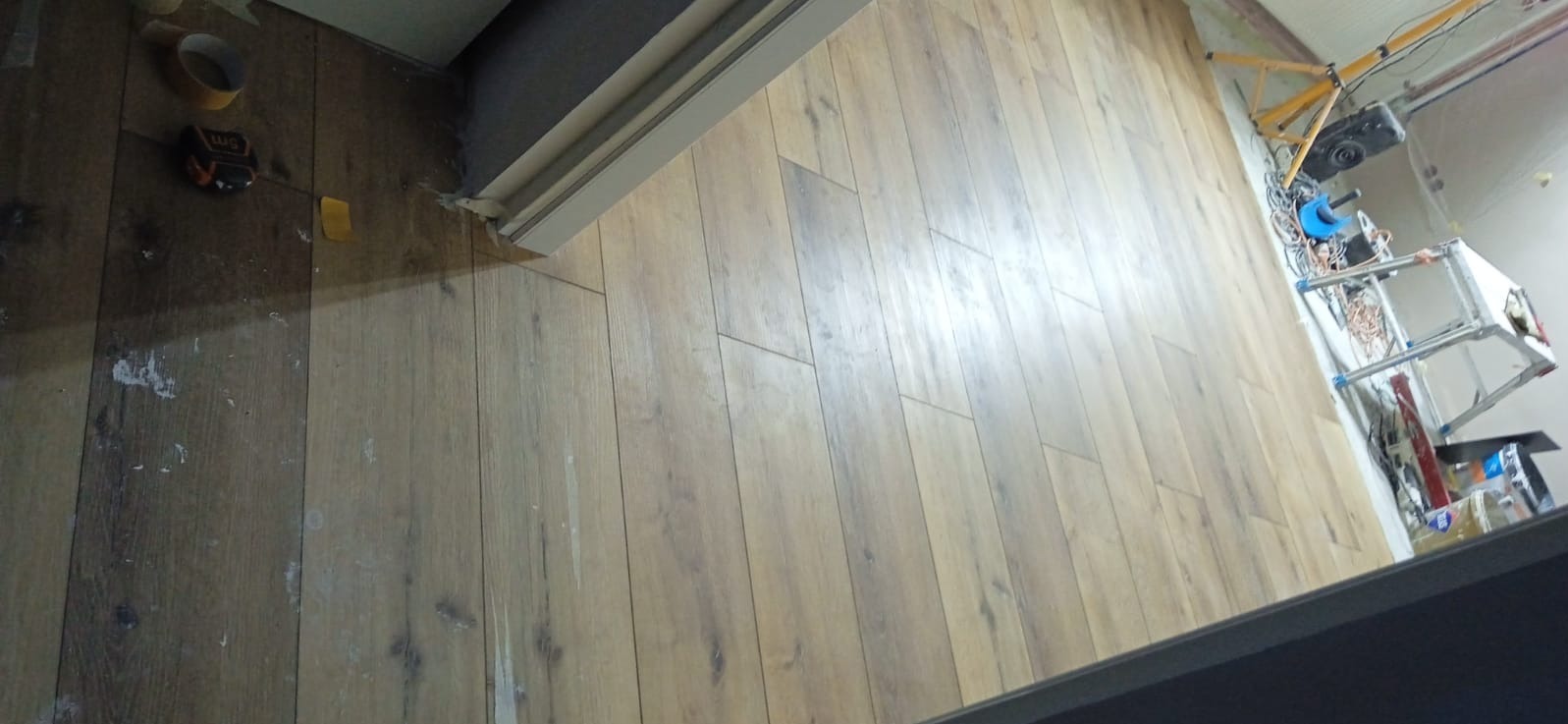 Ułożone panele podłogowe w trakcie prac wykończeniowych. Widoczne narzędzia, taśma miernicza i resztki po pracach. Ujęcie z góry, pod kątem.