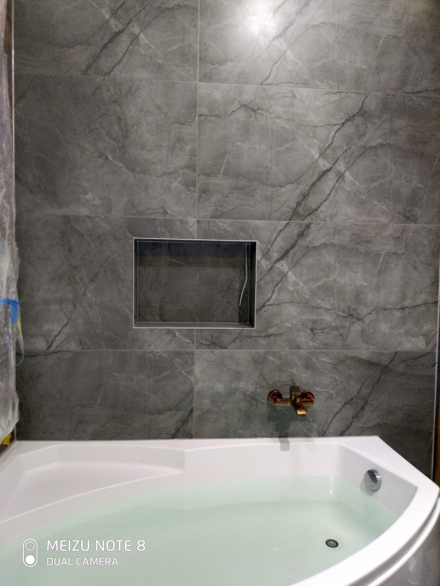 Wykończona łazienka z wanną i szarą, marmurkową glazurą. W ścianie wnęka, bateria w kolorze miedzi. Ujęcie z dołu, widoczny fragment wanny wypełnionej wodą.