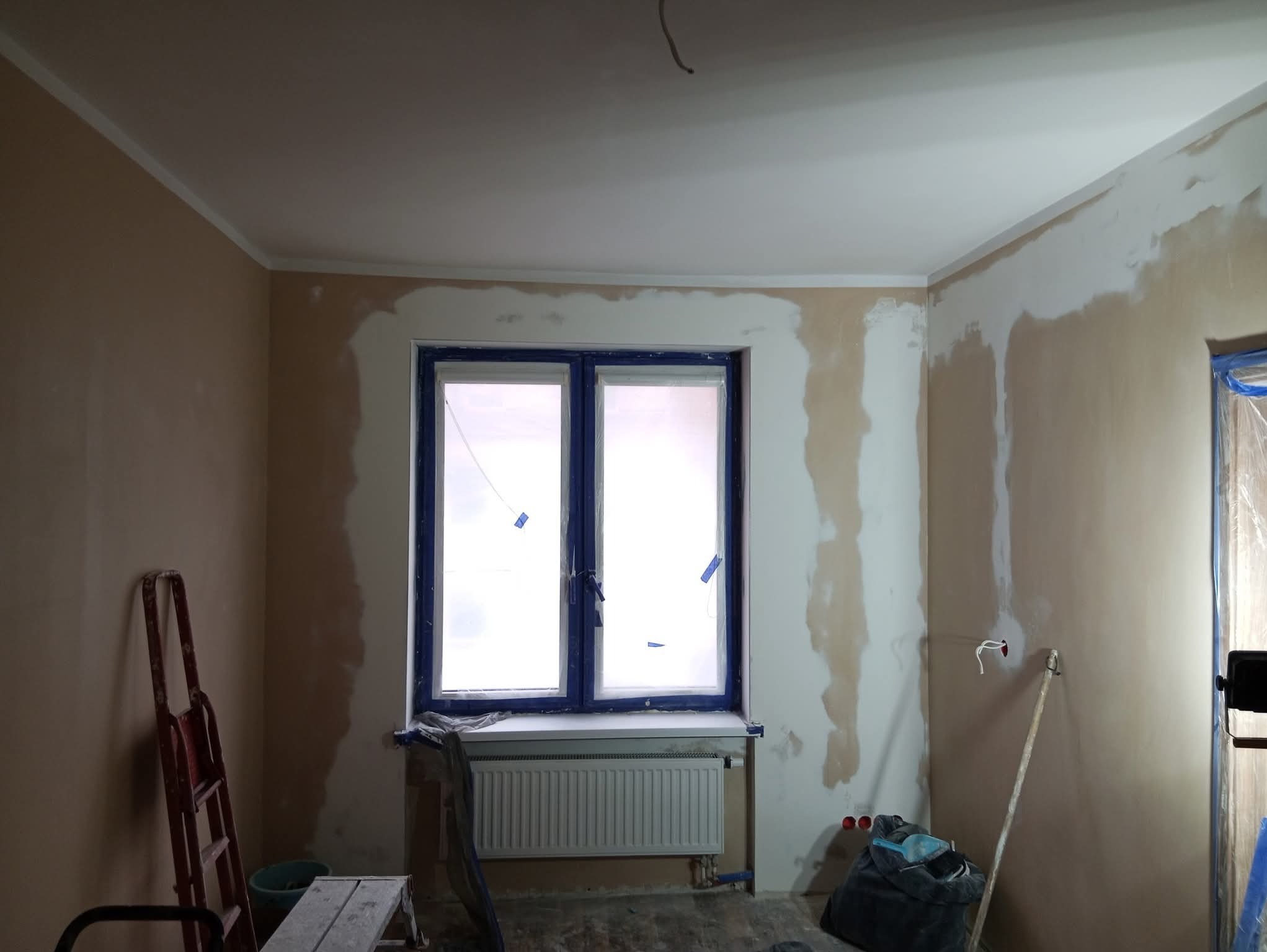 Pokój w trakcie szpachlowania ścian, okno zabezpieczone folią, widoczne narzędzia i drabina. Ściany częściowo pokryte gładzią, sufit pomalowany na biało.