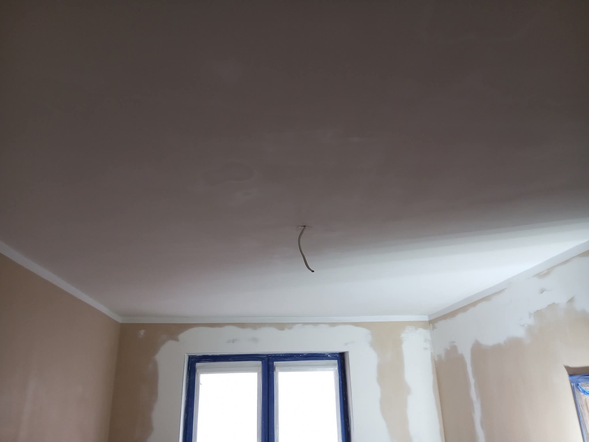 Pokój w trakcie prac wykończeniowych: gładzie na ścianach, pomalowany sufit z wystającym przewodem elektrycznym, okno zabezpieczone folią malarską.