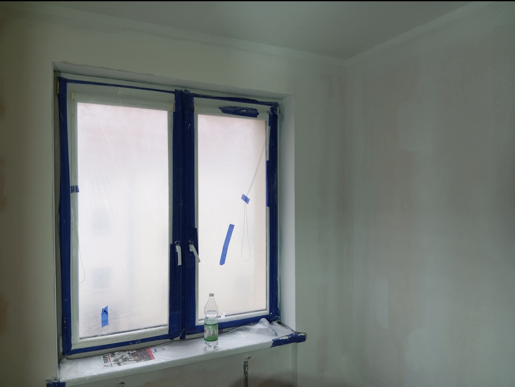 Pokój w trakcie przygotowań do malowania: okno zabezpieczone folią i taśmą, ściany po szpachlowaniu, widoczna butelka wody na parapecie. Przygotowanie do odświeżenia wnętrza.