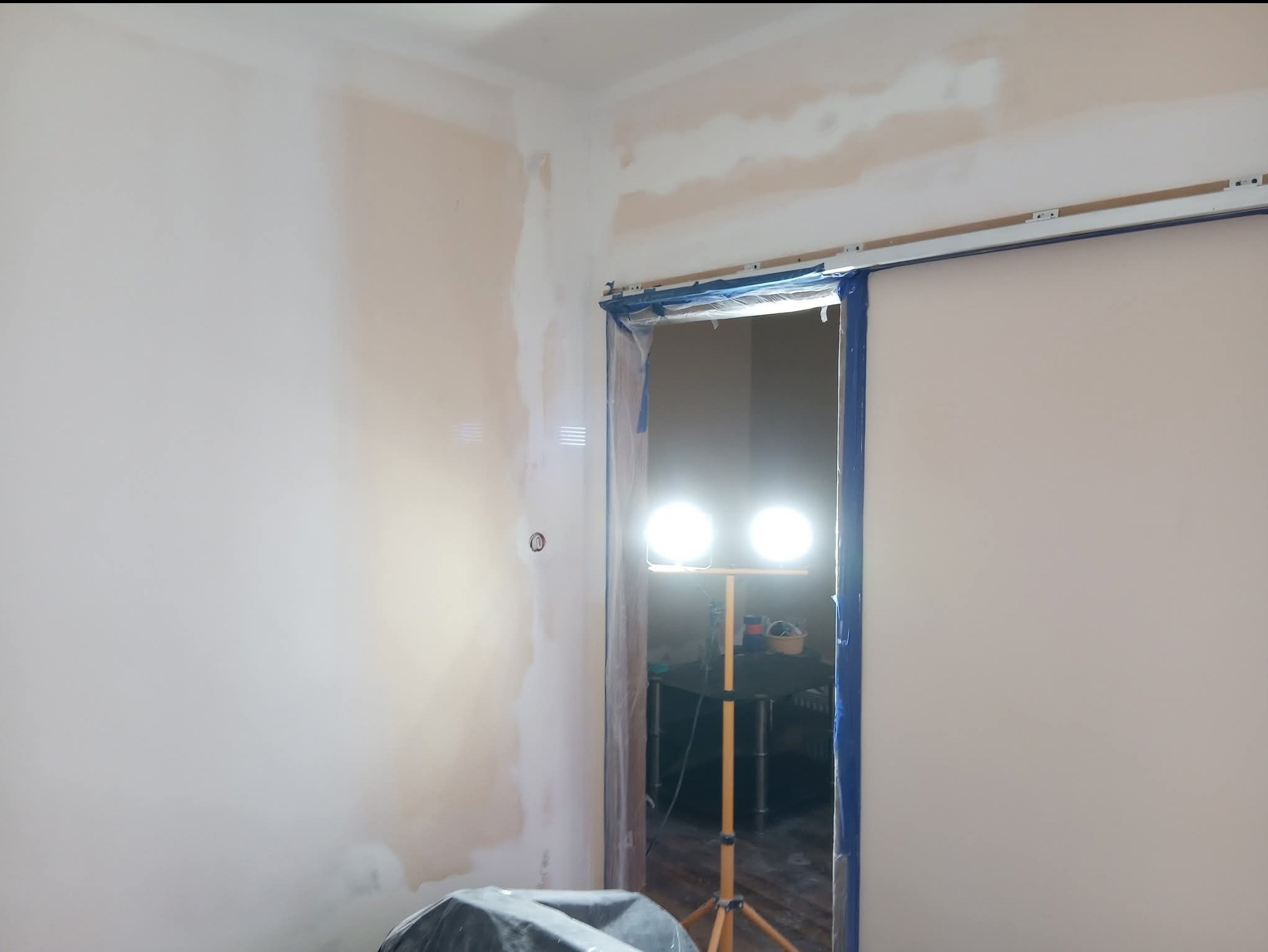 Ściana w trakcie szpachlowania, widoczne fragmenty nałożonej gładzi, zabezpieczone taśmą malarską futryny drzwiowe i lampa oświetlająca obszar roboczy.