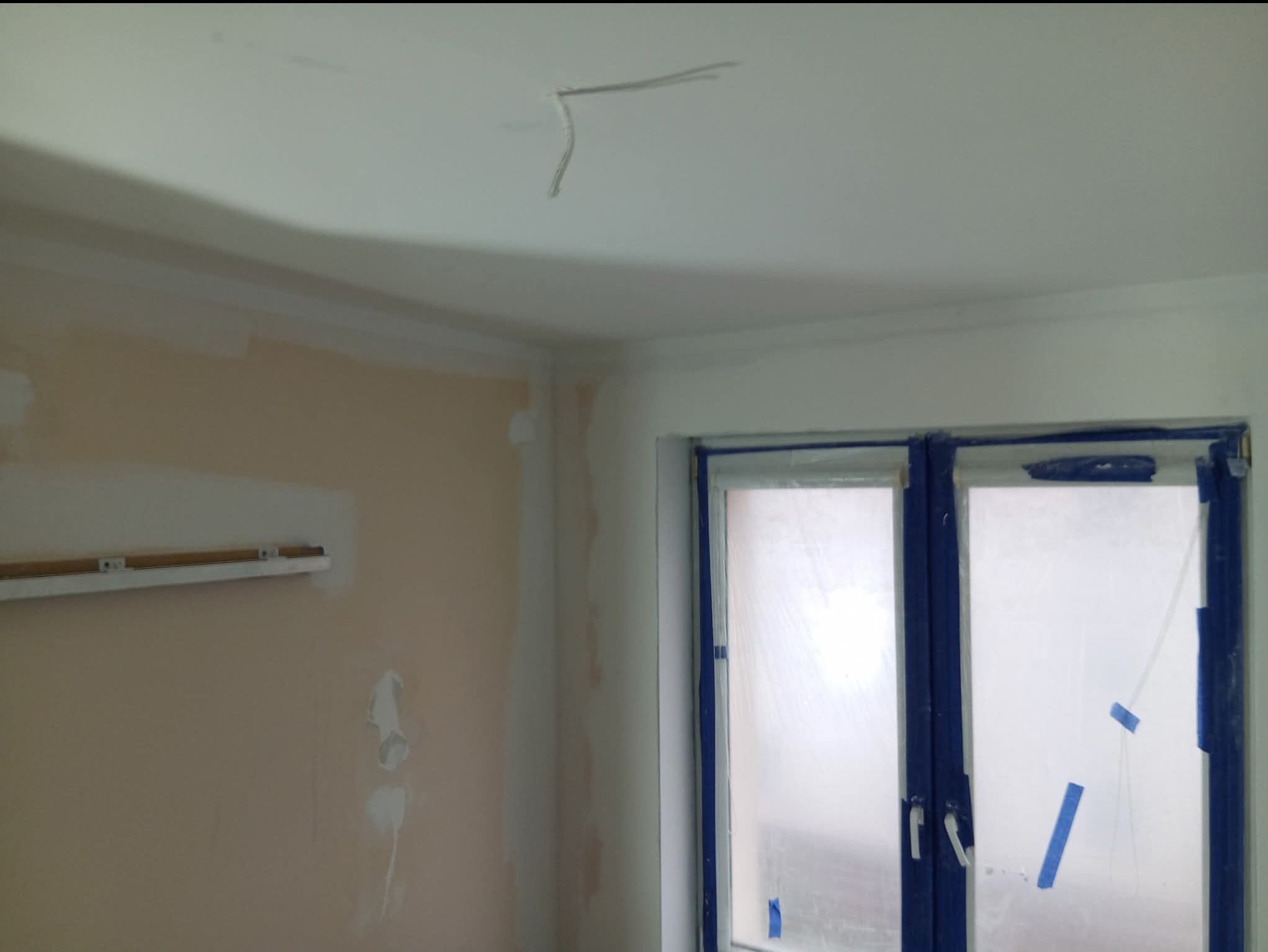 Pokój w trakcie remontu: ściany częściowo wyszpachlowane, okno zabezpieczone folią i taśmą malarską, widoczne wystające przewody elektryczne z sufitu.