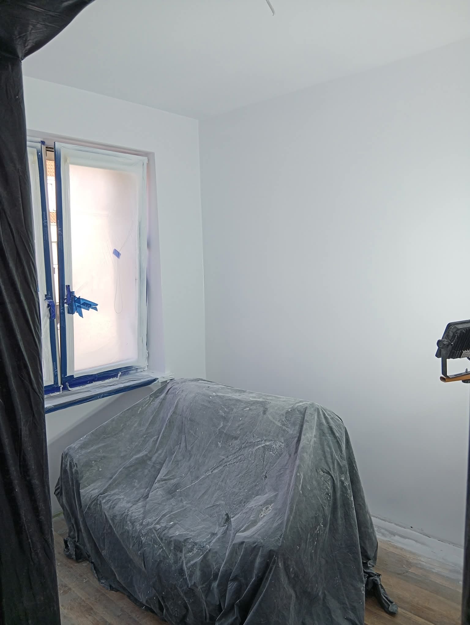 Pokój po malowaniu na biało, okno zabezpieczone folią, meble przykryte plandeką. Widoczne ślady farby na podłodze. Oświetlenie robocze w rogu.