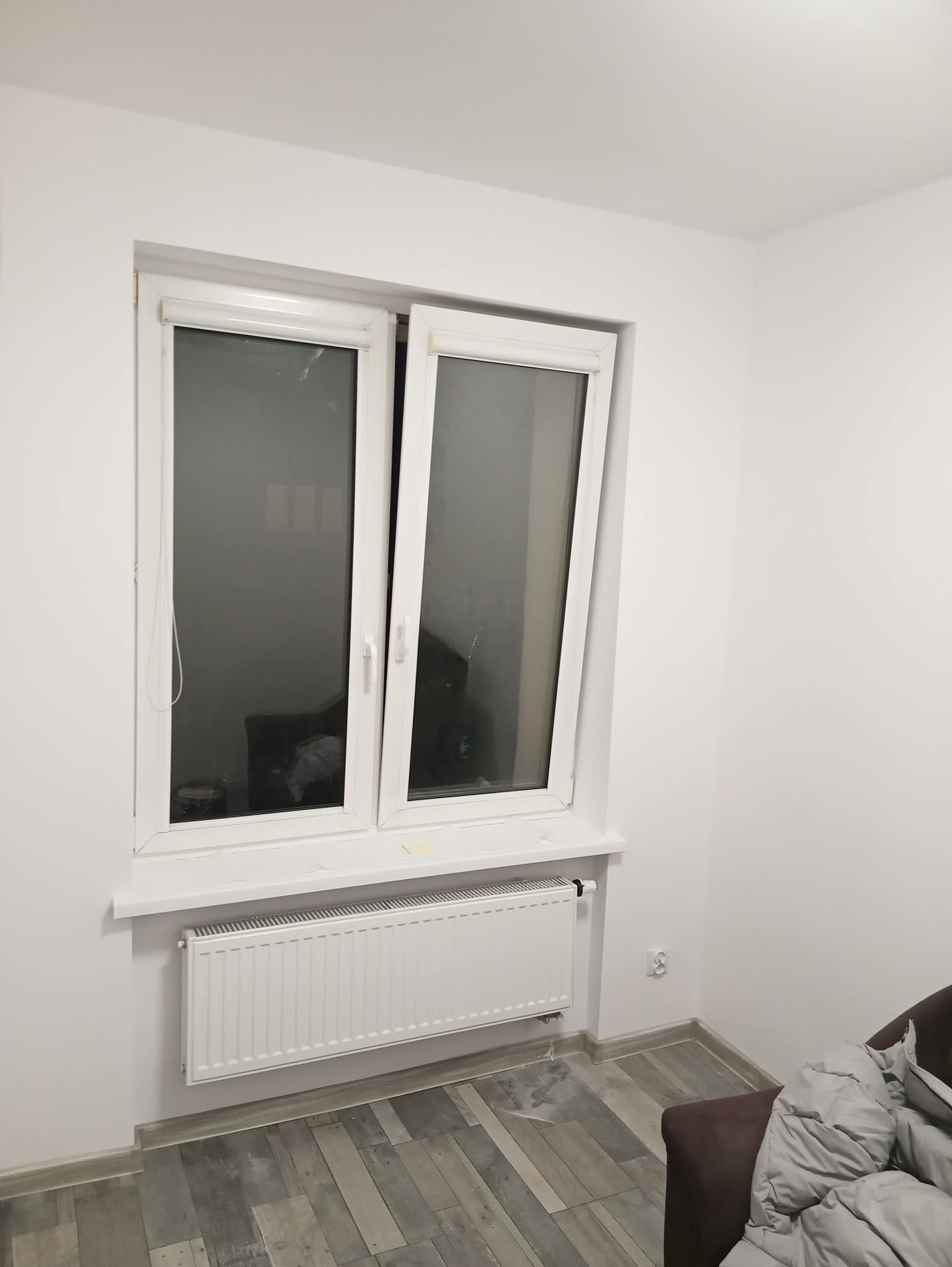 Białe, plastikowe okno uchylone w jasnym pokoju z grzejnikiem pod parapetem. Ściany pomalowane na biało, widoczny fragment podłogi i sofy.