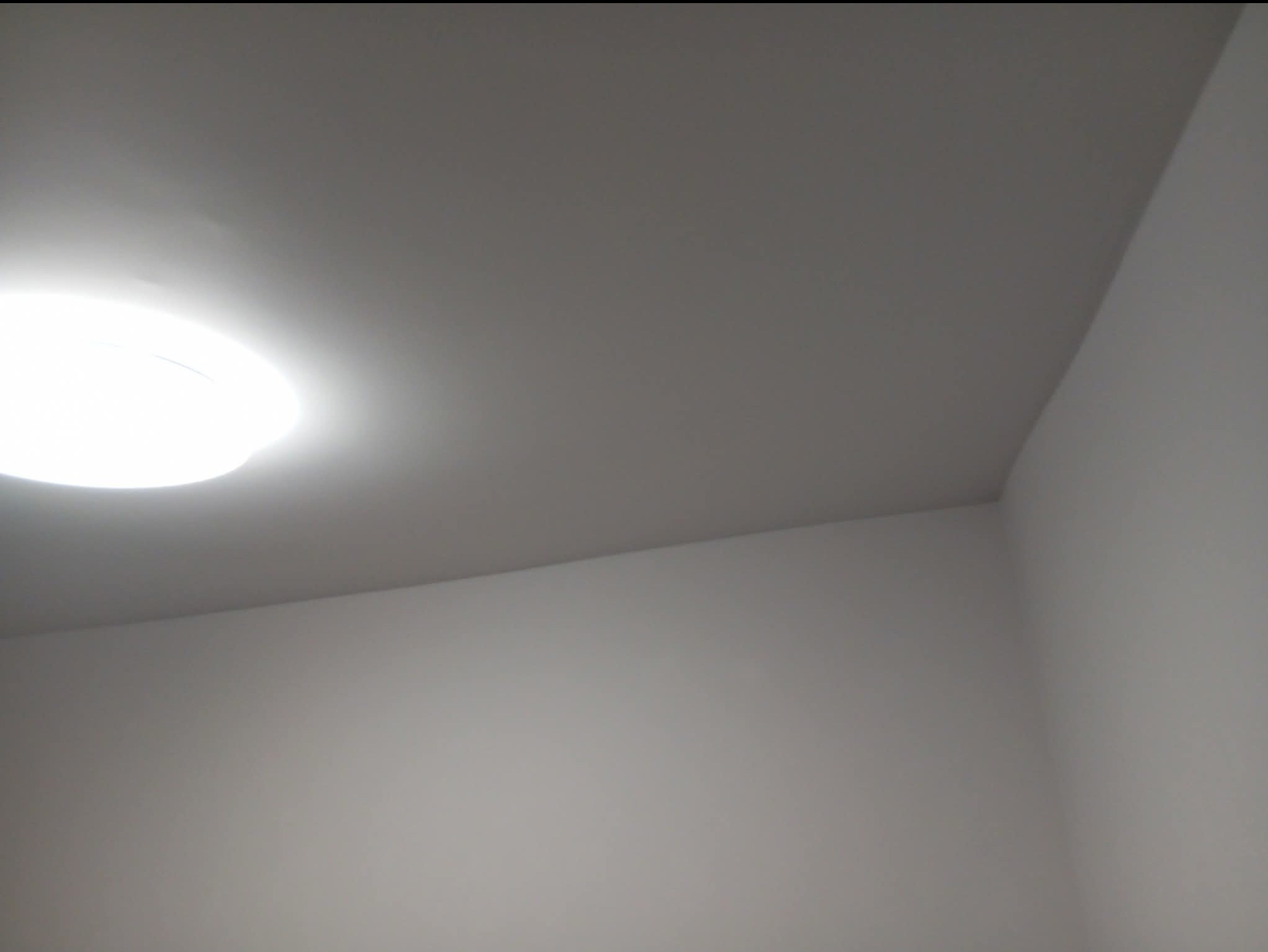 Gładka, pomalowana na szaro ściana w rogu pomieszczenia, oświetlona okrągłą lampą sufitową. Minimalistyczne wykończenie wnętrza.