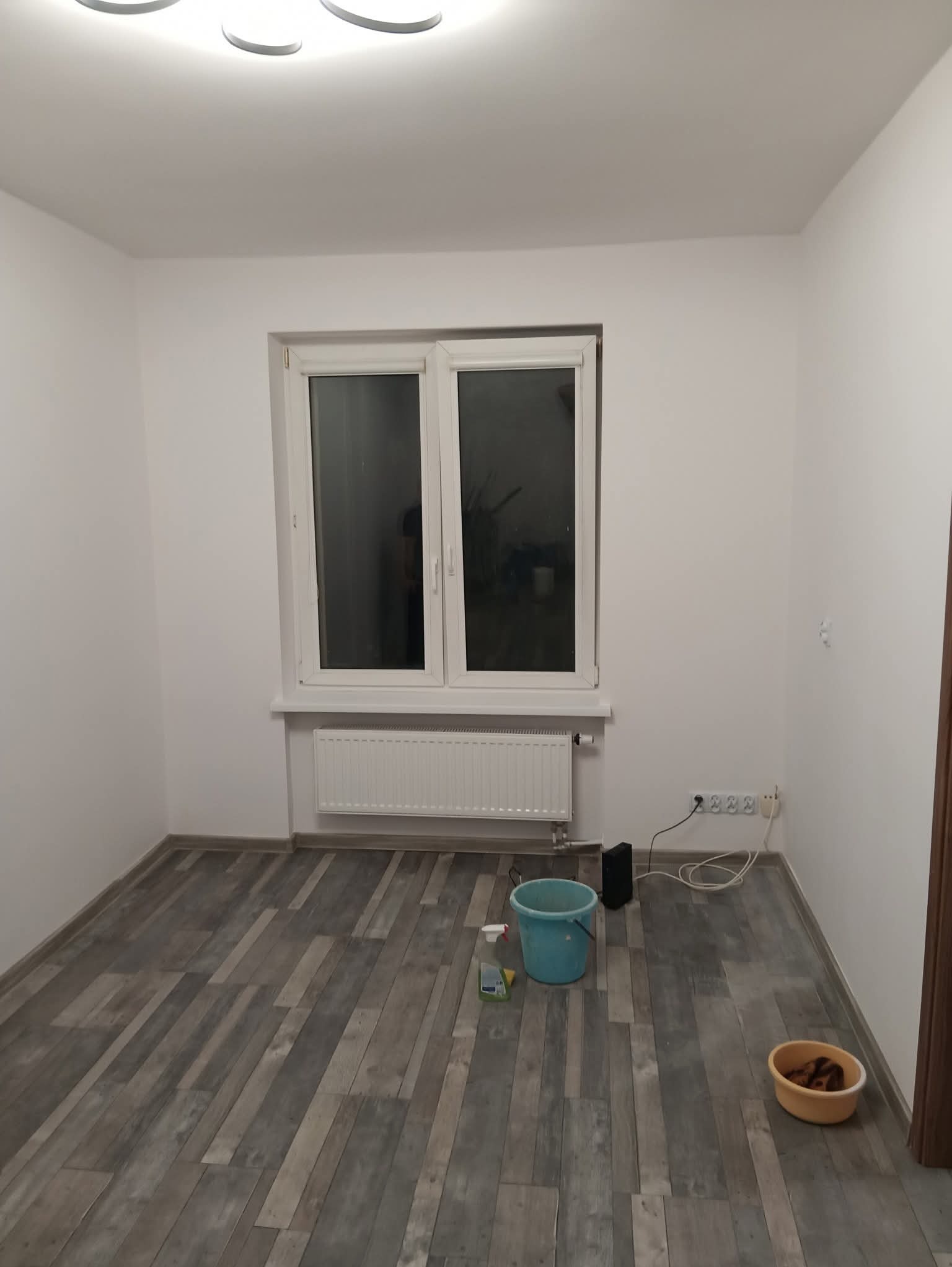 Pusty pokój po odświeżeniu: białe ściany, szara podłoga imitująca drewno, okno z grzejnikiem. Widoczne akcesoria do sprzątania: wiadro i miska.