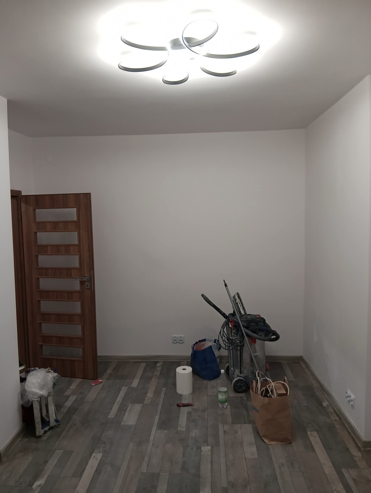 Pokój przed remontem: białe ściany, drewniana podłoga, narzędzia malarskie i szlifierka pod ścianą. Widoczna lampa sufitowa i drzwi z przeszkleniami.