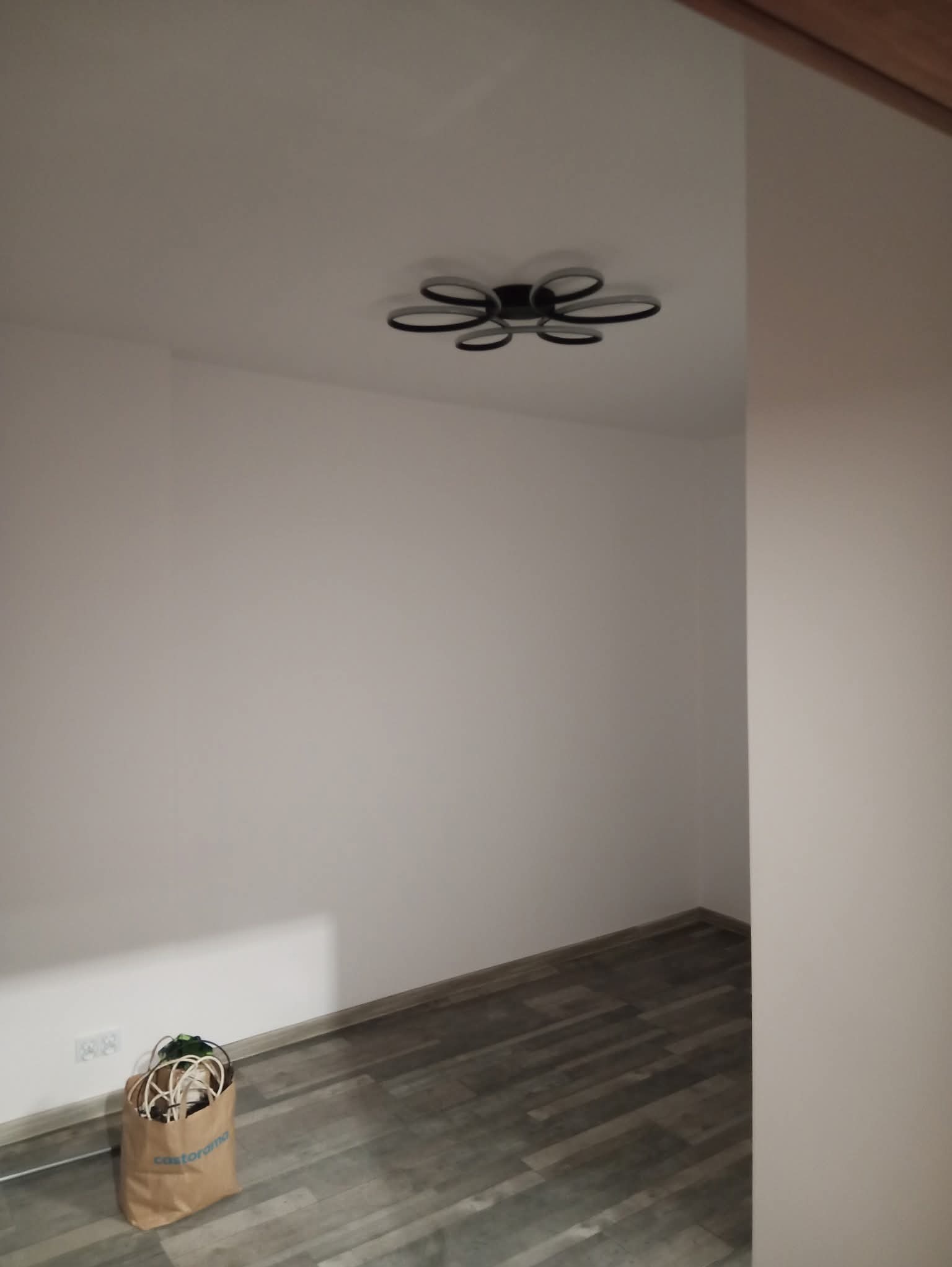 Pusty pokój z szarą podłogą, białe ściany i nowoczesna lampa sufitowa. W rogu torba Castorama. Minimalistyczne wnętrze przed wykończeniem.