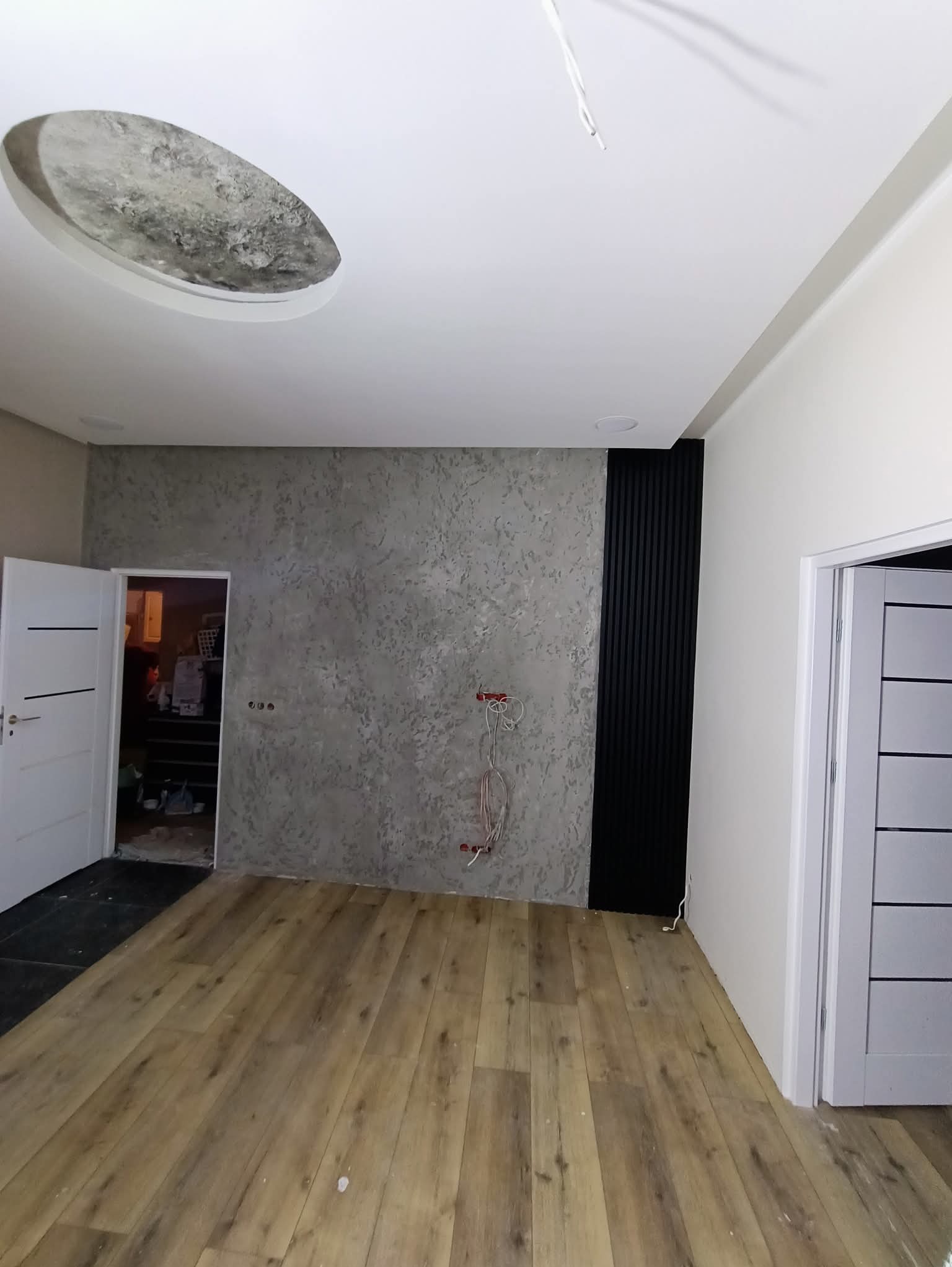 Pokój po wykończeniu: drewnopodobna podłoga, ściana z szarą fakturą, czarne panele dekoracyjne, białe drzwi i sufit z okrągłym wgłębieniem. Widoczne kable elektryczne.