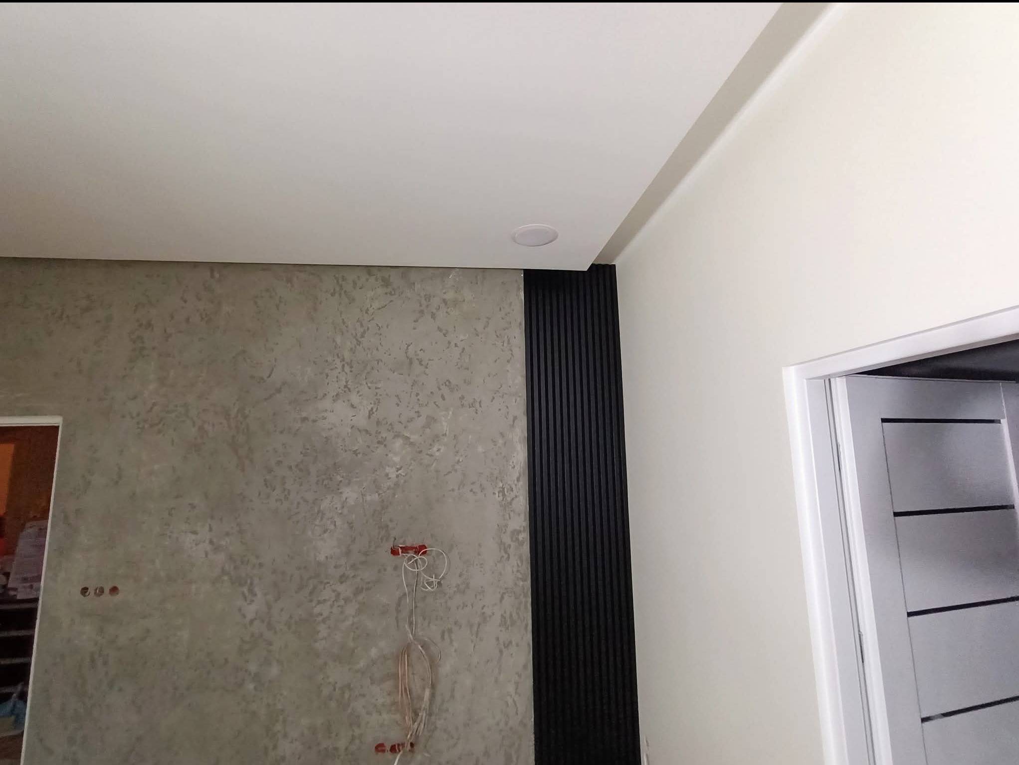 Wykończona ściana z dekoracyjną strukturą i panelem pionowym, widoczne gniazdka elektryczne przed montażem osprzętu, fragment sufitu i drzwi.