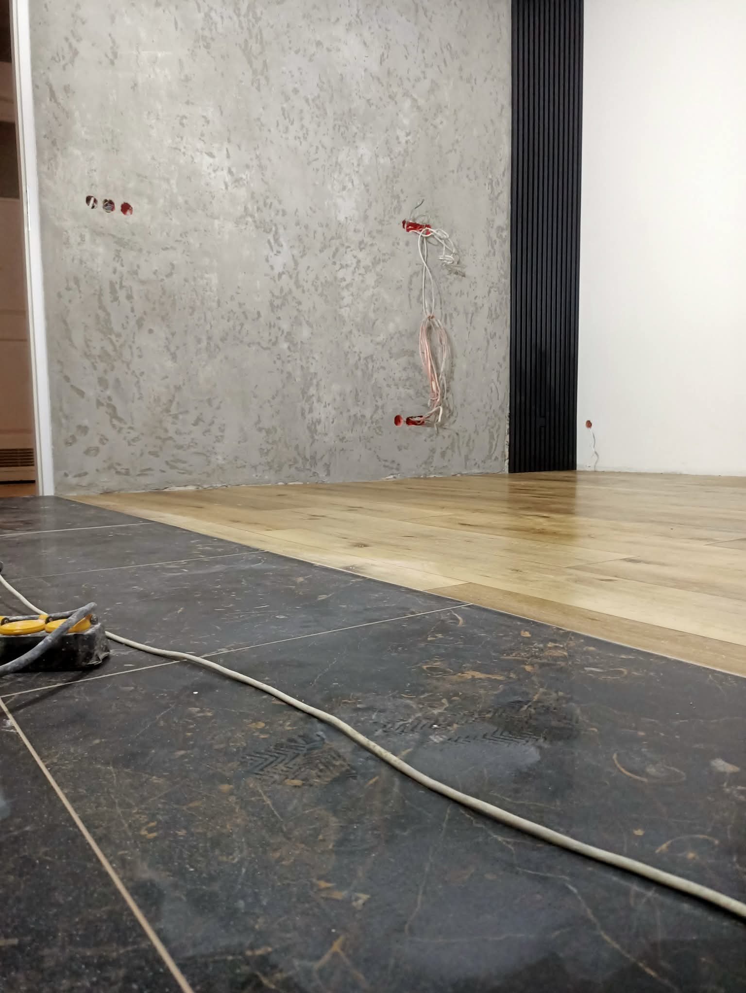 Wnętrze w trakcie prac: połączenie szarej ściany z dekoracyjną fakturą, czarnych paneli ściennych i jasnej drewnianej podłogi. Widoczne kable elektryczne i narzędzia.