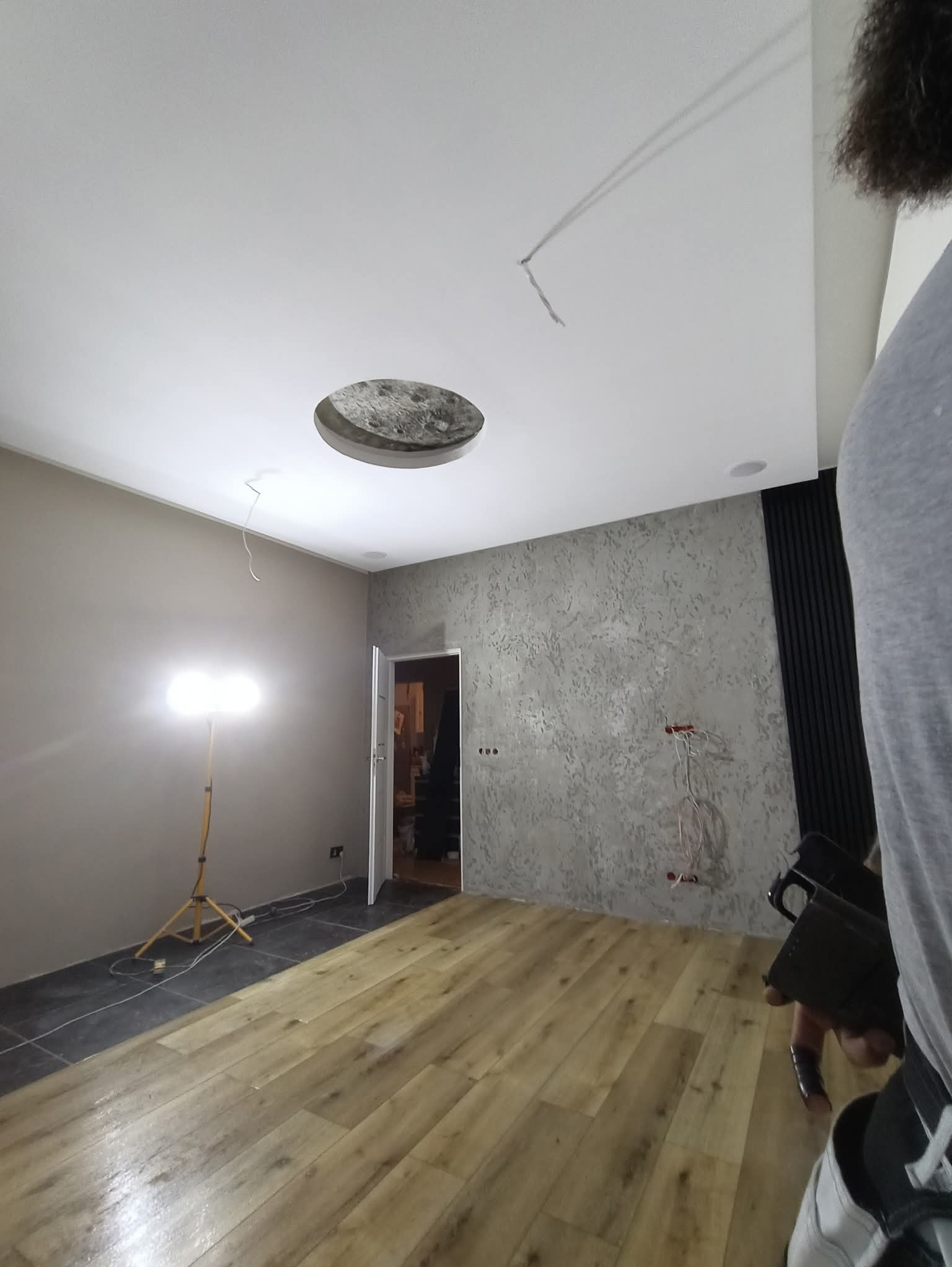 Wykończone wnętrze z drewnianą podłogą, szarą ścianą z fakturą i białym sufitem z otworem na oświetlenie. Widoczna lampa na statywie i fragment osoby.