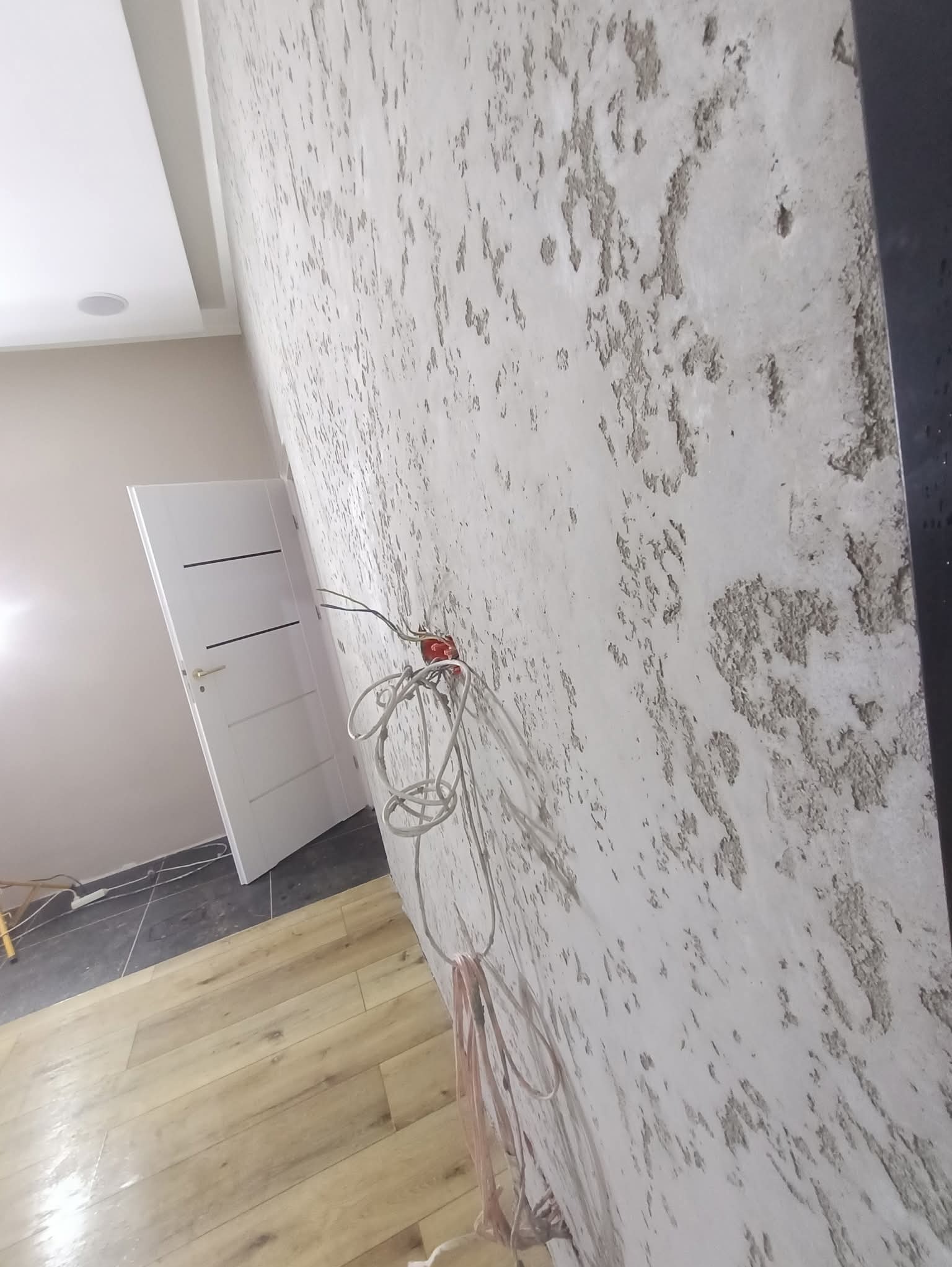 Teksturowana ściana w odcieniach szarości, z widocznym gniazdkiem elektrycznym i przewodami. W tle białe drzwi i fragment podłogi z paneli.