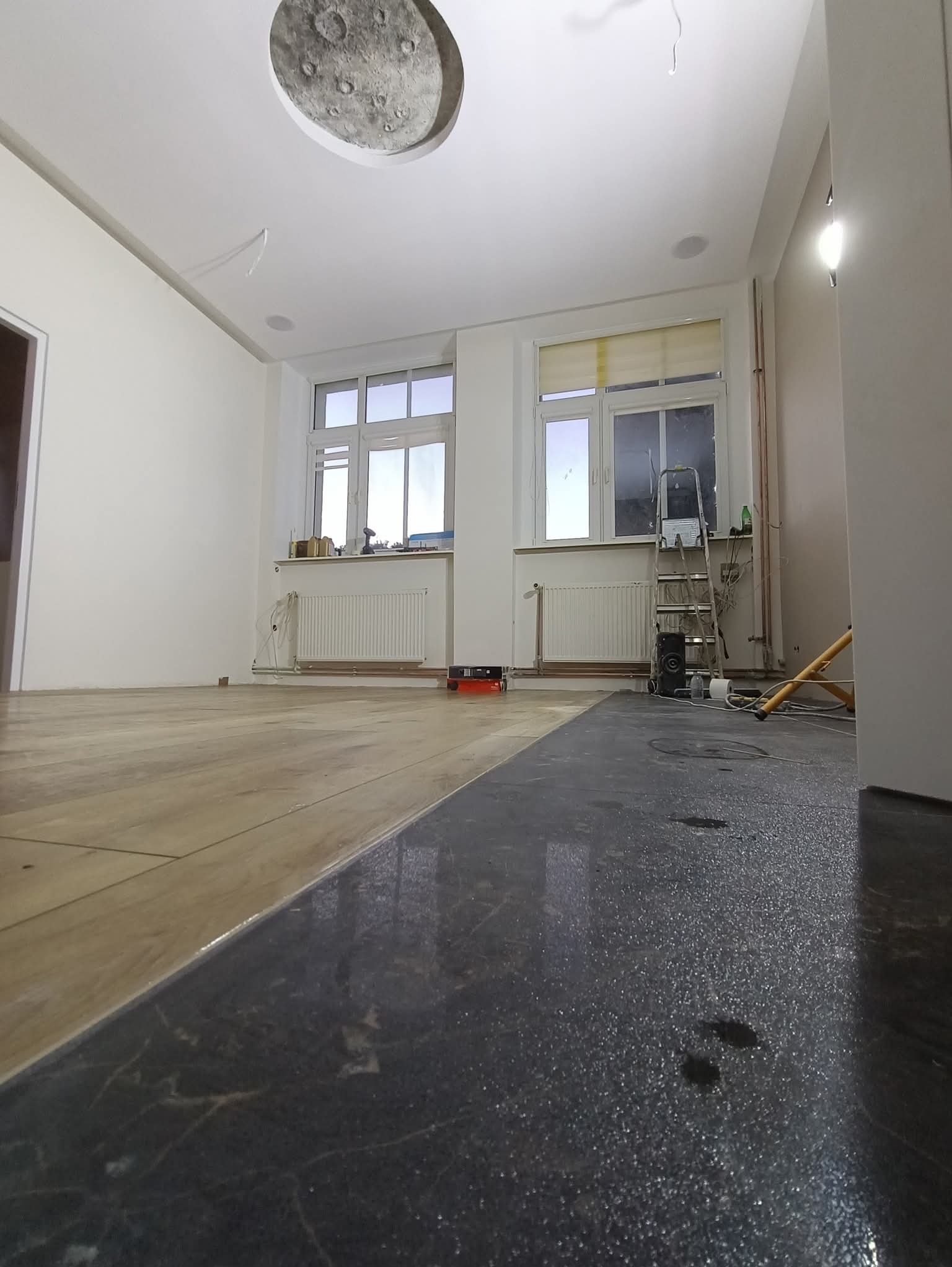 Pokój po częściowym położeniu podłogi: panele drewniane i ciemne płytki. Widoczna drabina, okna z grzejnikami i sufit z dekoracją imitującą księżyc.