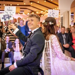 PetermanFoto - Bartosz Peterman - Uśmiechnięty pan młody trzyma kartę z napisem 'Ja' podczas wesela. Panna młoda w koronkowej sukni siedzi obok, trzymając kartę 'Ona'. W tle goście weselni.