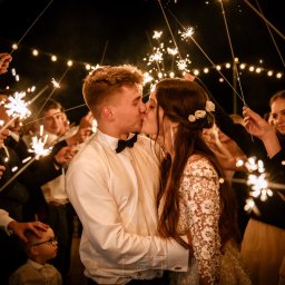 PetermanFoto - Bartosz Peterman - Pocałunek pary młodej otoczonej gośćmi z zimnymi ogniami, ciepłe światło lampek w tle, uroczysta atmosfera, suknia z koronką, czarna mucha, chłopiec w okularach.