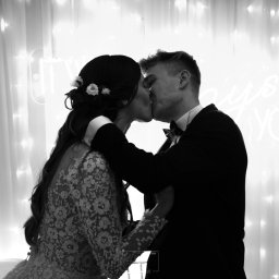 PetermanFoto - Bartosz Peterman - Czarno-białe zdjęcie całującej się pary młodej na tle białych zasłon i neonowego napisu. Panna młoda ma kwiaty we włosach i koronkową suknię.