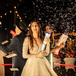 PetermanFoto - Bartosz Peterman - Uśmiechnięta panna młoda w koronkowej sukni, otoczona bańkami mydlanymi i strumieniem banknotów z pistoletów na tle dekoracji świetlnych i gości.