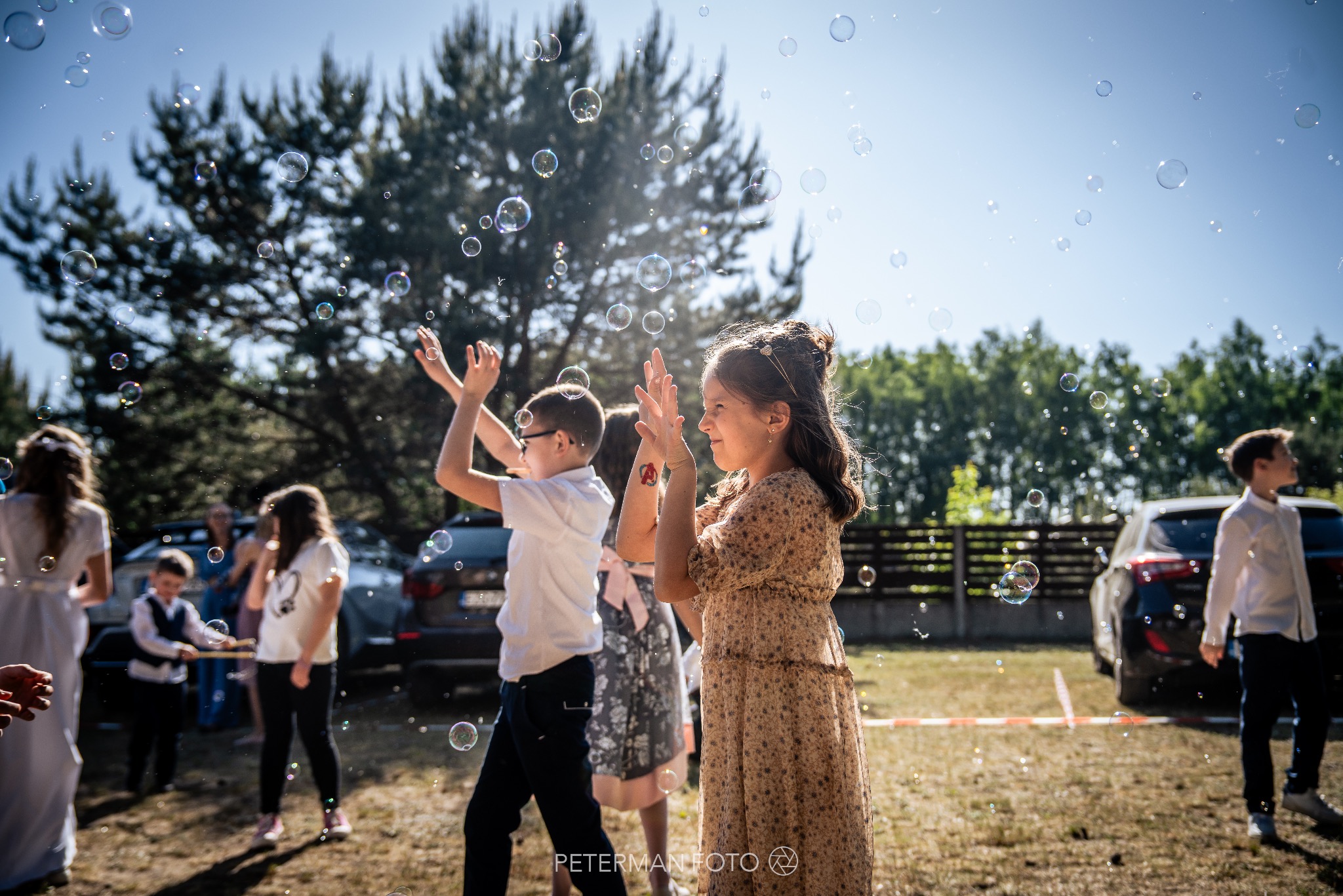Dzieci w plenerze bawią się w otoczeniu baniek mydlanych, w tle drzewa i samochody. Dziewczynka z ozdobą we włosach i tatuażem na ręce. Jasne, słoneczne światło.