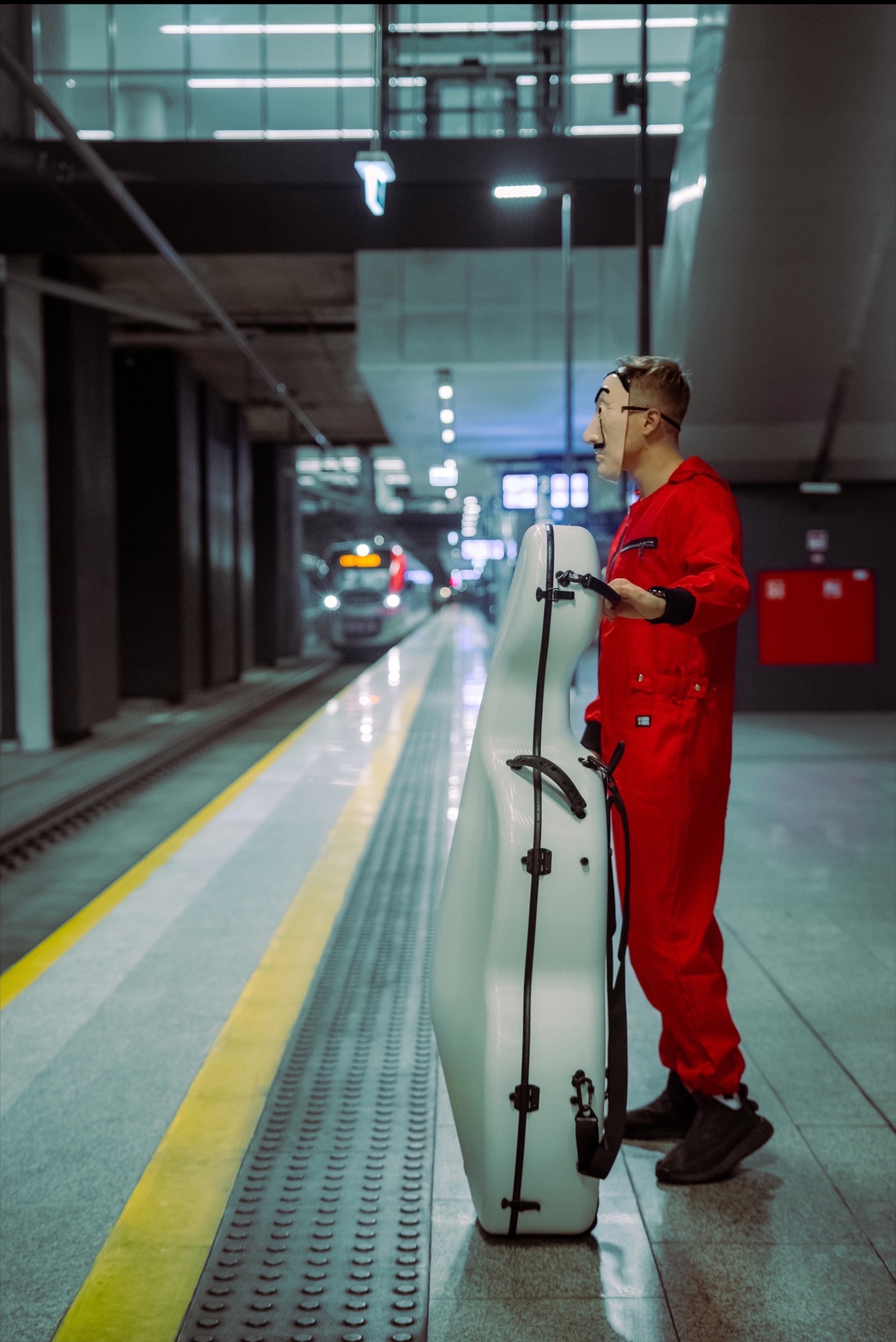 Osoba w czerwonym kombinezonie i masce Dalego czeka na peronie metra z futerałem na kontrabas. W tle rozmyty pociąg.