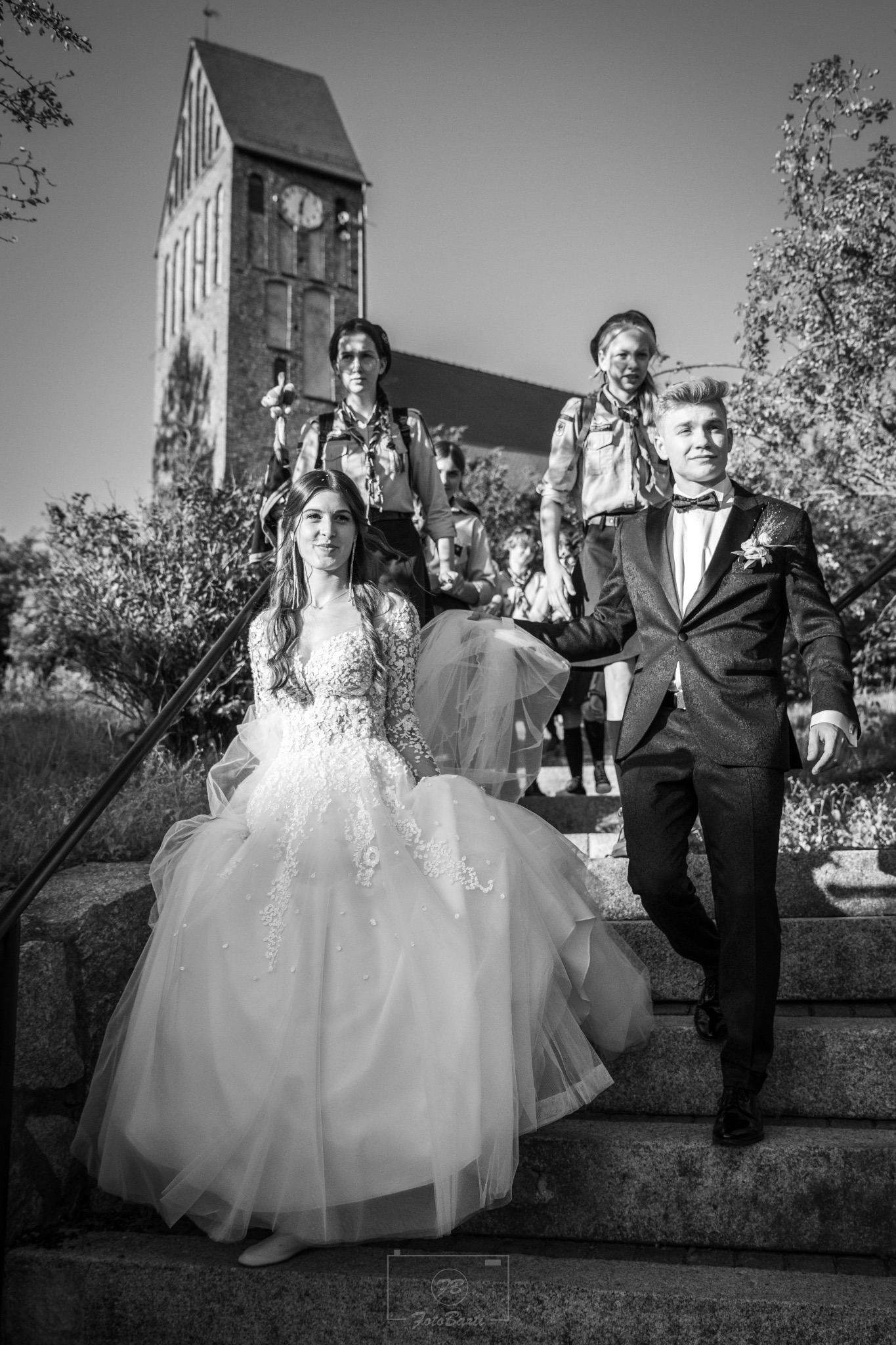 Czarno-białe zdjęcie pary młodej schodzącej po kamiennych schodach, za nimi grupa skautów, w tle wieża kościoła z zegarem. Ujęcie z dołu.
