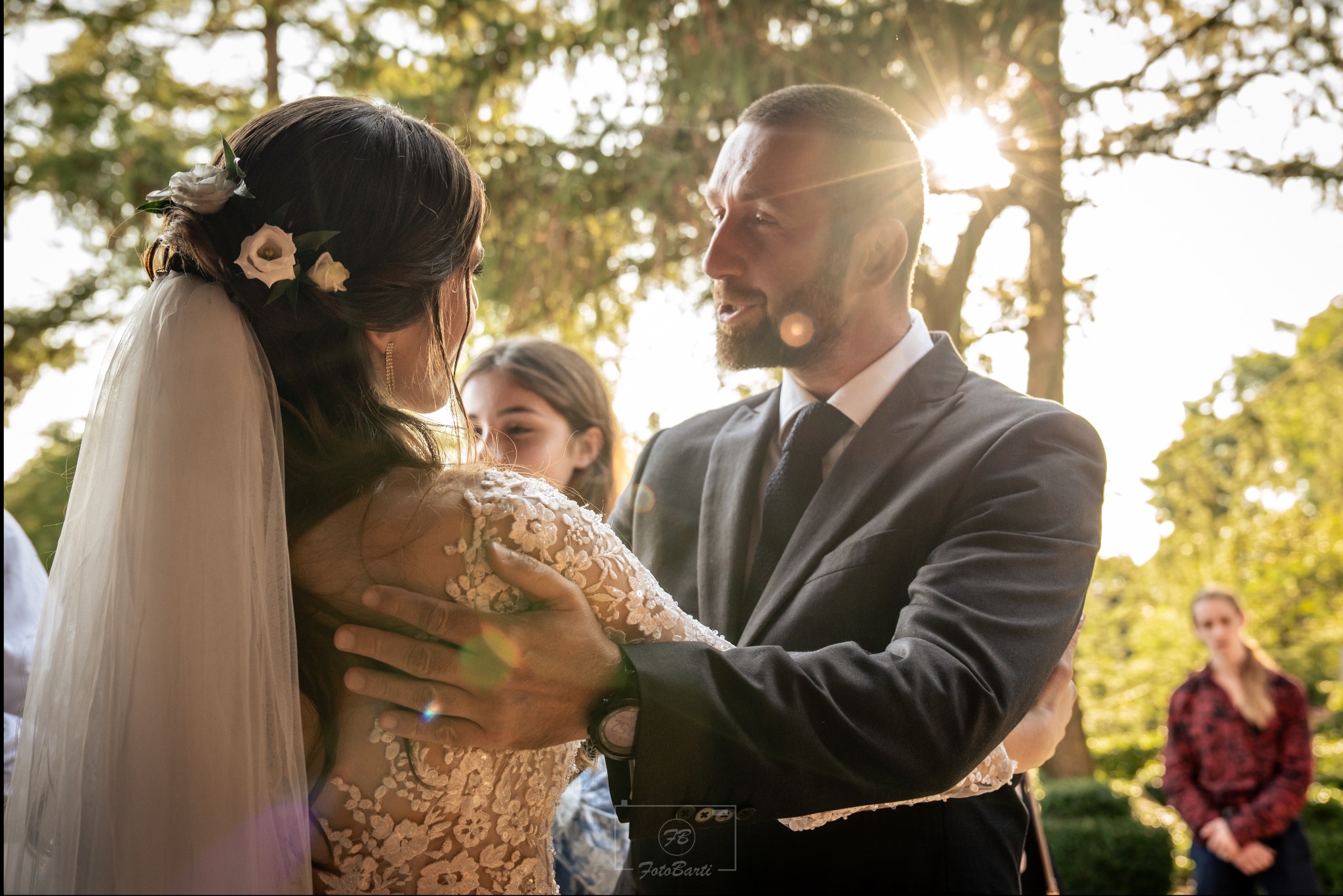 Para młoda w objęciach podczas słonecznego dnia. Panna młoda w sukni z koronkowymi rękawami i welonem, pan młody w garniturze. W tle zieleń, rozproszone światło.
