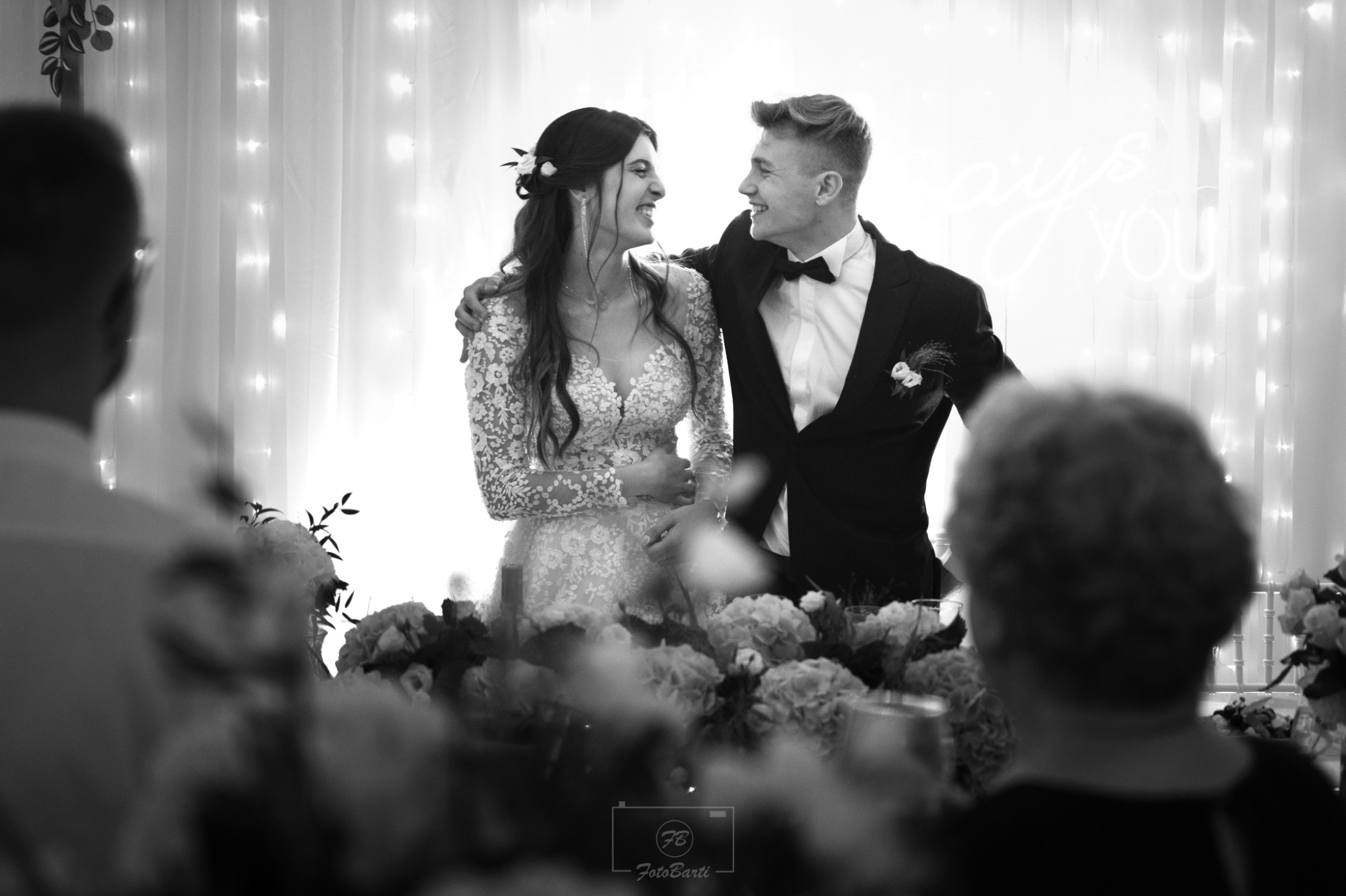 Czarno-białe zdjęcie uśmiechniętej pary młodej podczas wesela, z dekoracją kwiatową na pierwszym planie i neonowym napisem w tle. Pan młody w smokingu z muchą.