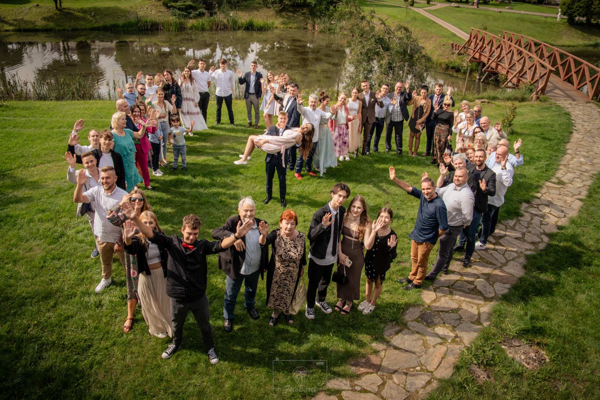 Duża grupa ludzi w plenerze, ustawiona w kształcie serca, macha do kamery. Mężczyzna trzyma kobietę na rękach. W tle widać staw i drewniany mostek.