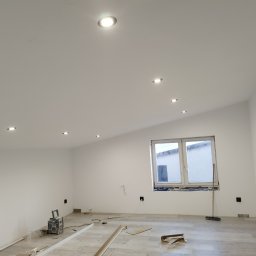Artur Jagielski - Białe wnętrze po odświeżeniu: ściany, sufit z punktowym oświetleniem, okno. Na podłodze panele i narzędzia, sugerujące prace wykończeniowe.
