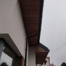 Artur Jagielski - Fragment elewacji budynku z drewnianą podbitką i czarną rynną. Widoczne okno z drewnianą ramą i fragment sąsiedniego budynku w tle.