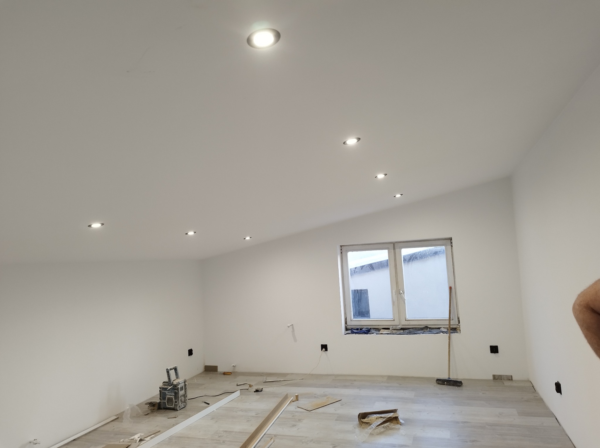 Białe wnętrze po odświeżeniu: ściany, sufit z punktowym oświetleniem, okno. Na podłodze panele i narzędzia, sugerujące prace wykończeniowe.