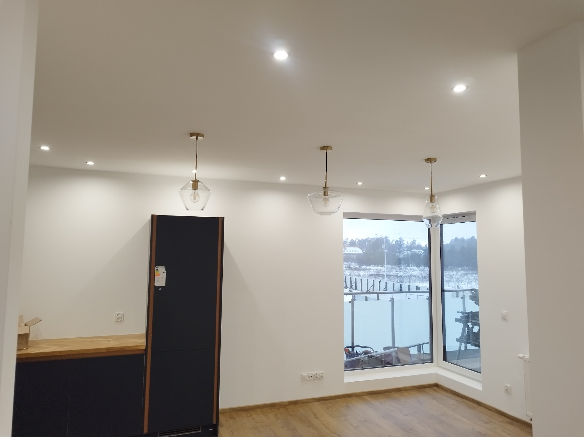 Nowoczesna kuchnia z czarnymi szafkami, drewnianym blatem i trzema szklanymi lampami wiszącymi. Widok na zewnątrz przez duże okno.