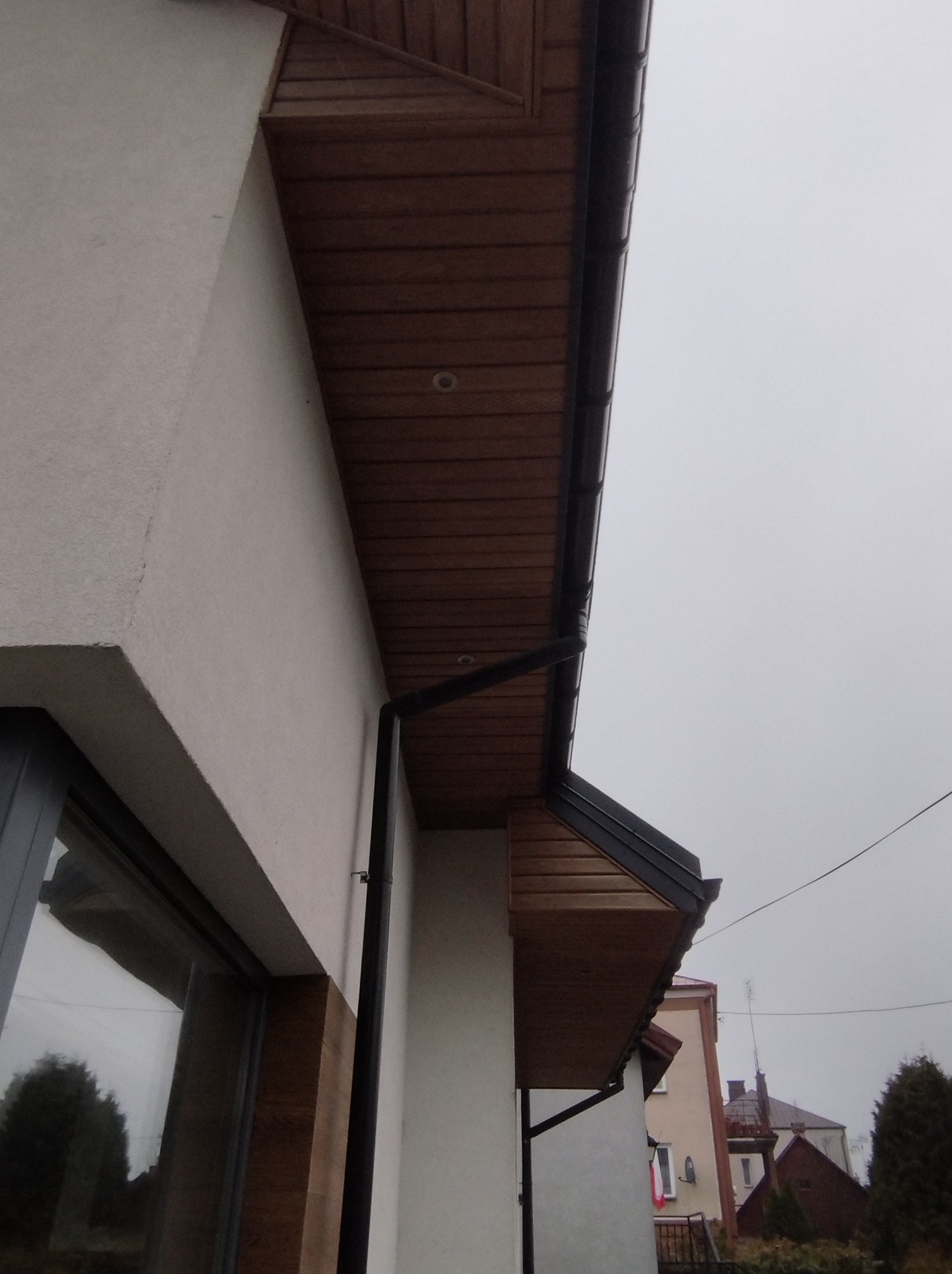Fragment elewacji budynku z drewnianą podbitką i czarną rynną. Widoczne okno z drewnianą ramą i fragment sąsiedniego budynku w tle.