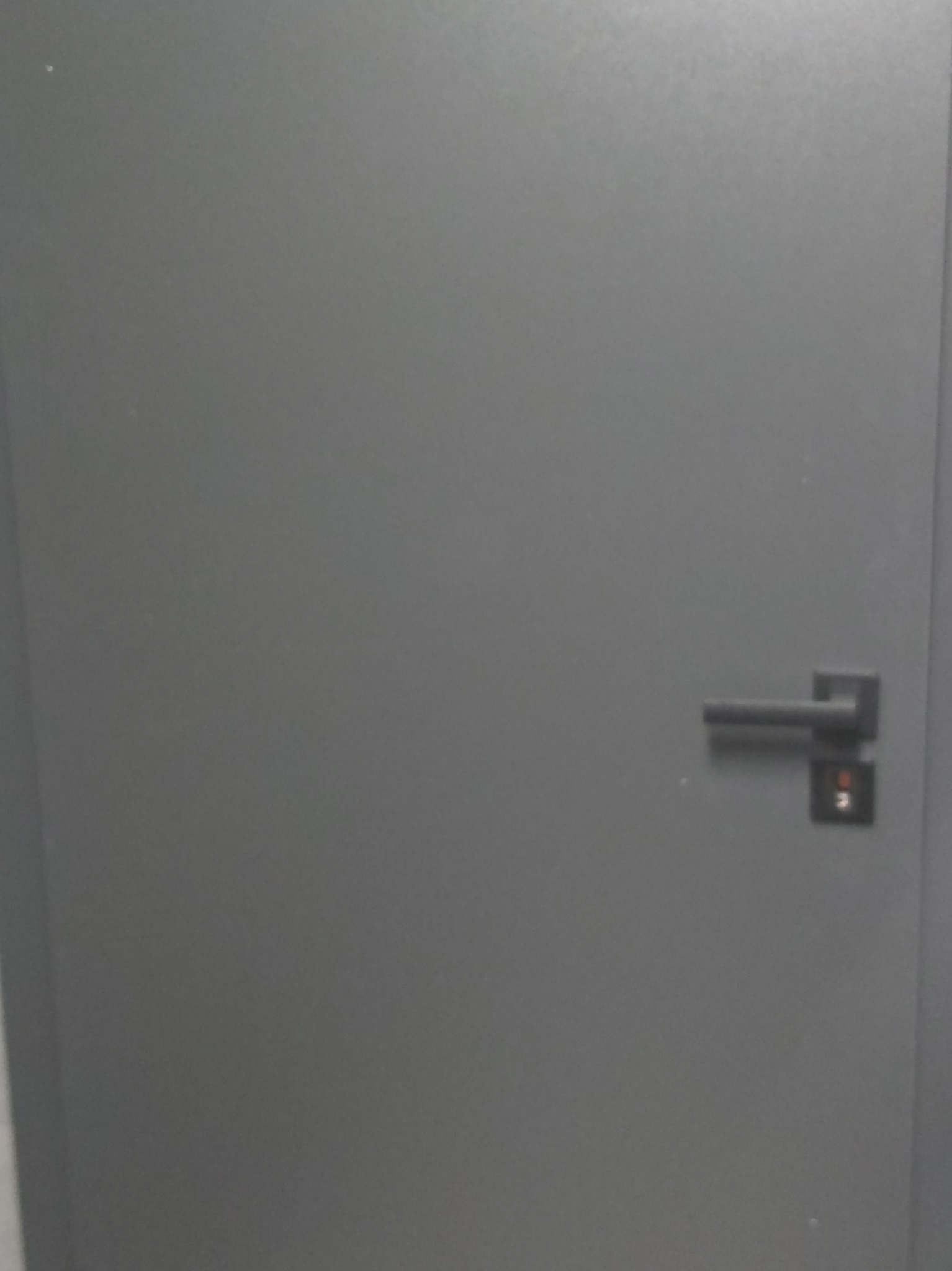 Szare, gładkie drzwi wewnętrzne z czarną klamką i zamkiem. Minimalistyczny design, jednolite wykończenie, widoczny fragment ościeżnicy.