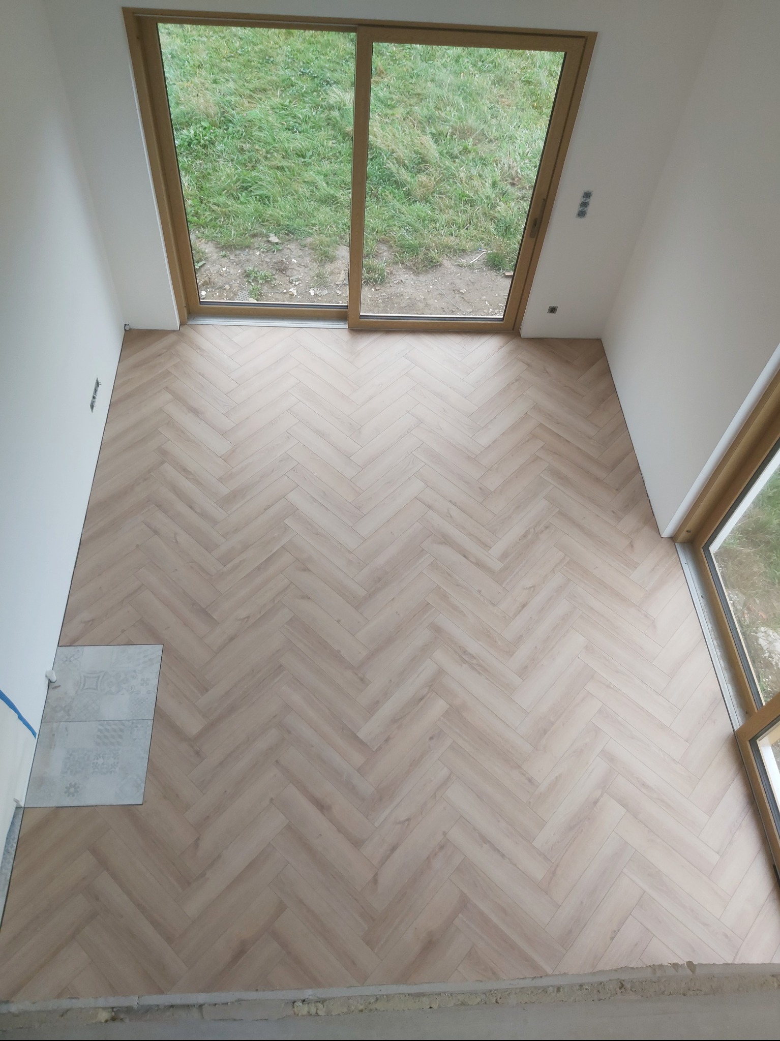 Jasne panele ułożone w jodełkę w nowym pokoju z dużymi oknami. Fragment płytek podłogowych w rogu. Widok z góry na świeżo wykończoną podłogę.