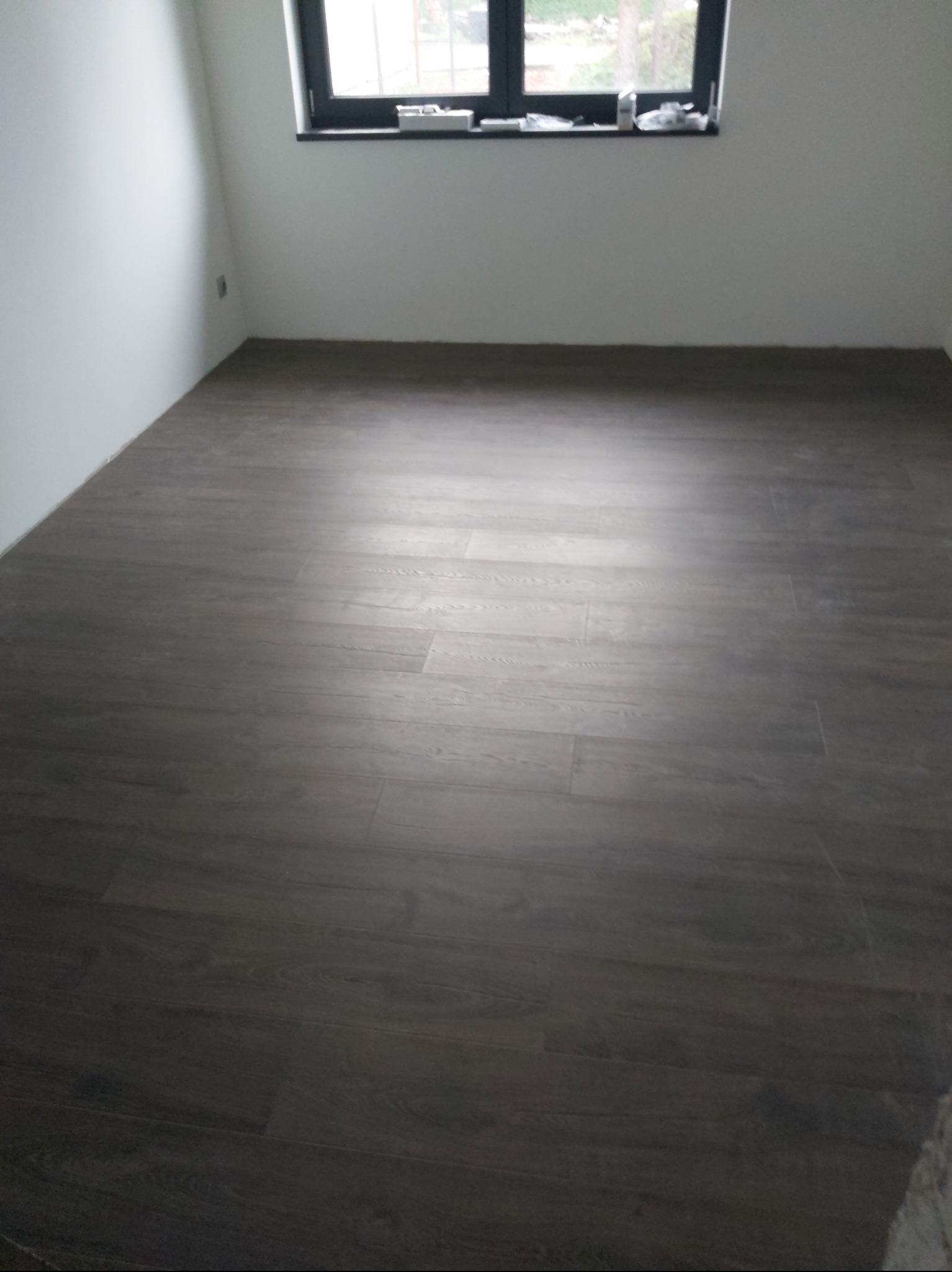 Ułożone panele podłogowe w pokoju z oknem. Ciemne panele imitujące drewno, białe ściany. Widok na całą podłogę, równo ułożone panele, brak listew przypodłogowych.