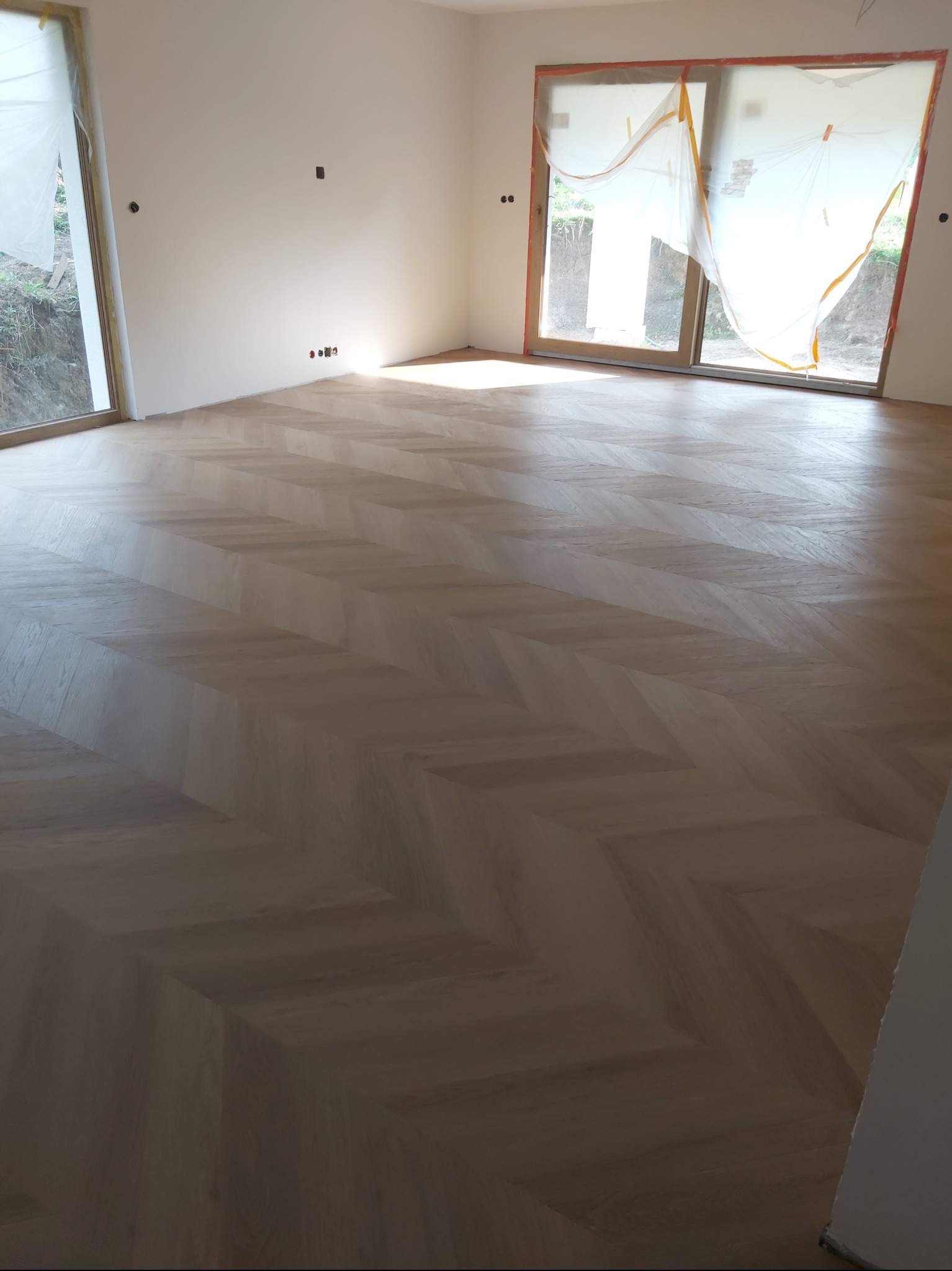 Jasny parkiet ułożony we wzór jodełki francuskiej w przestronnym, słonecznym pokoju z dużymi oknami zabezpieczonymi folią malarską. Widoczne gniazdka elektryczne.