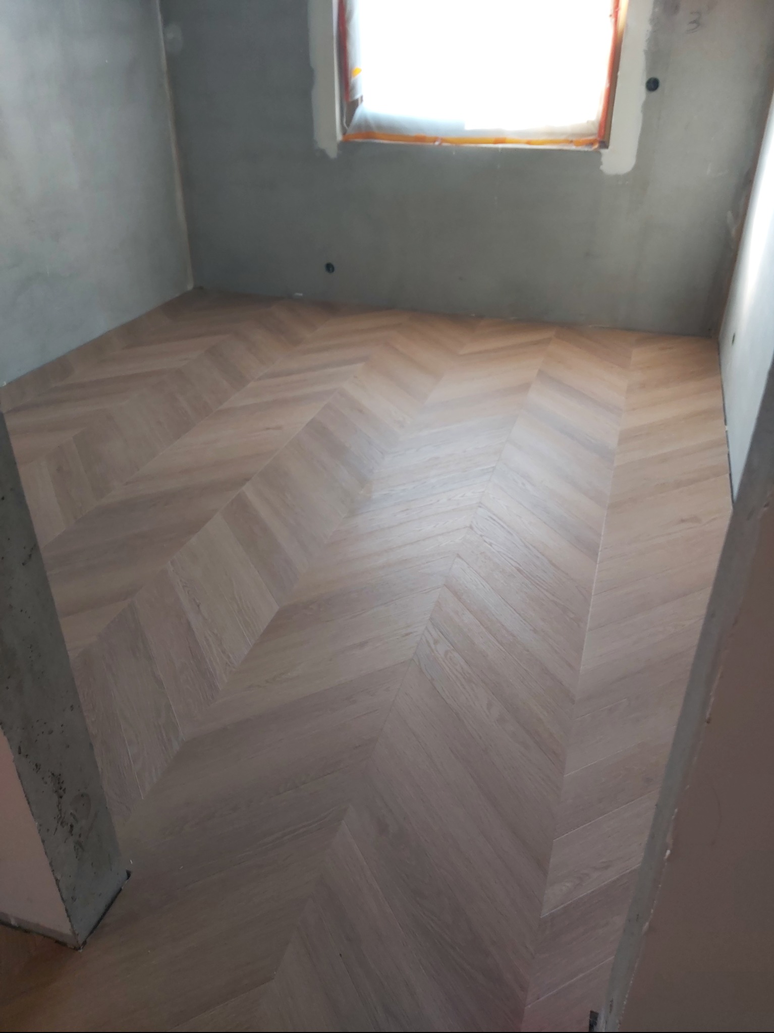 Jasne panele ułożone w jodełkę w surowym wnętrzu z oknem. Widoczne ślady po pracach wykończeniowych na ścianach. Charakterystyczny wzór podłogi.