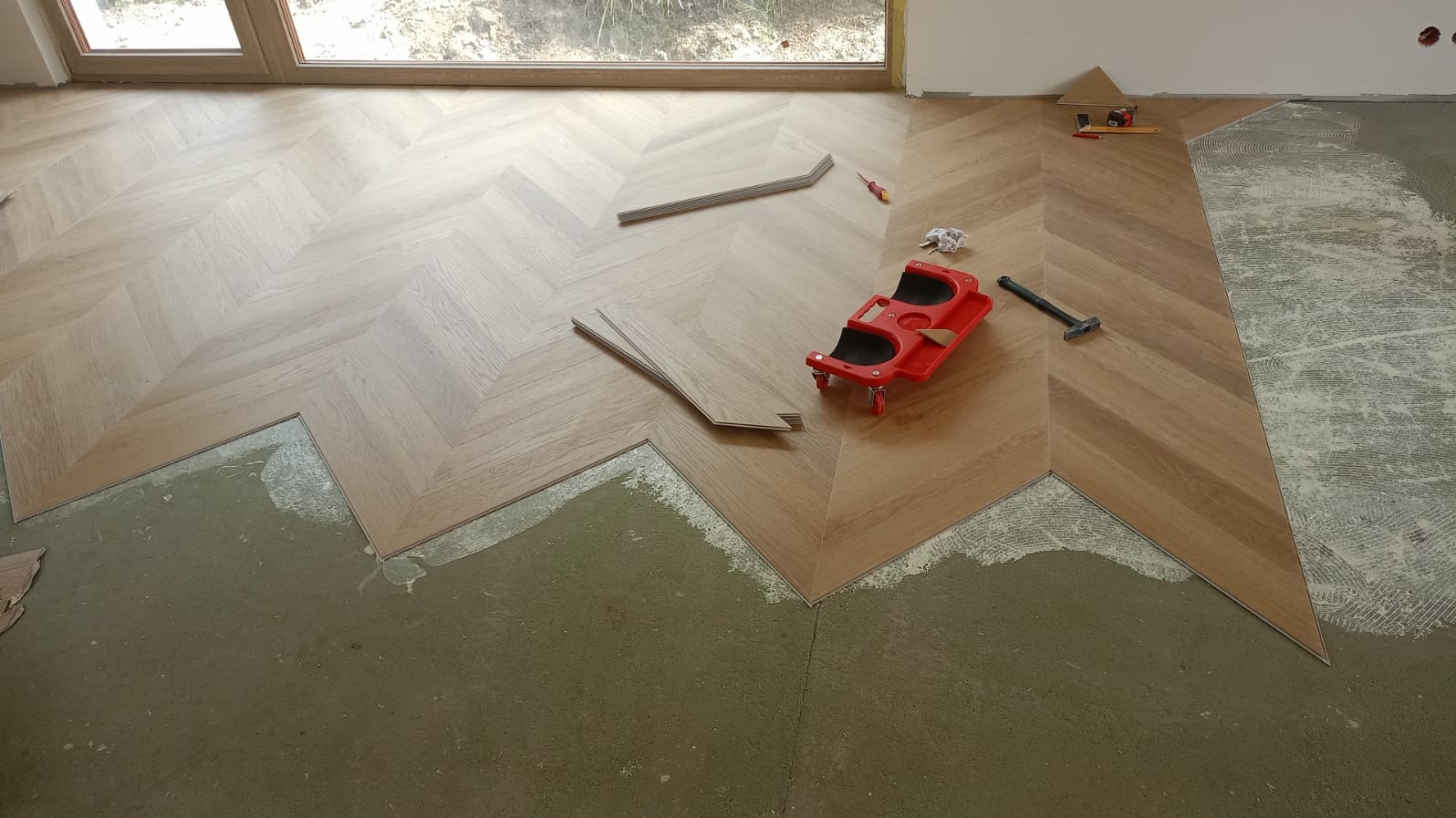 Układanie jasnych paneli podłogowych w jodełkę francuską. Widoczne narzędzia i fragment podłoża przed położeniem. Częściowo ułożona podłoga.