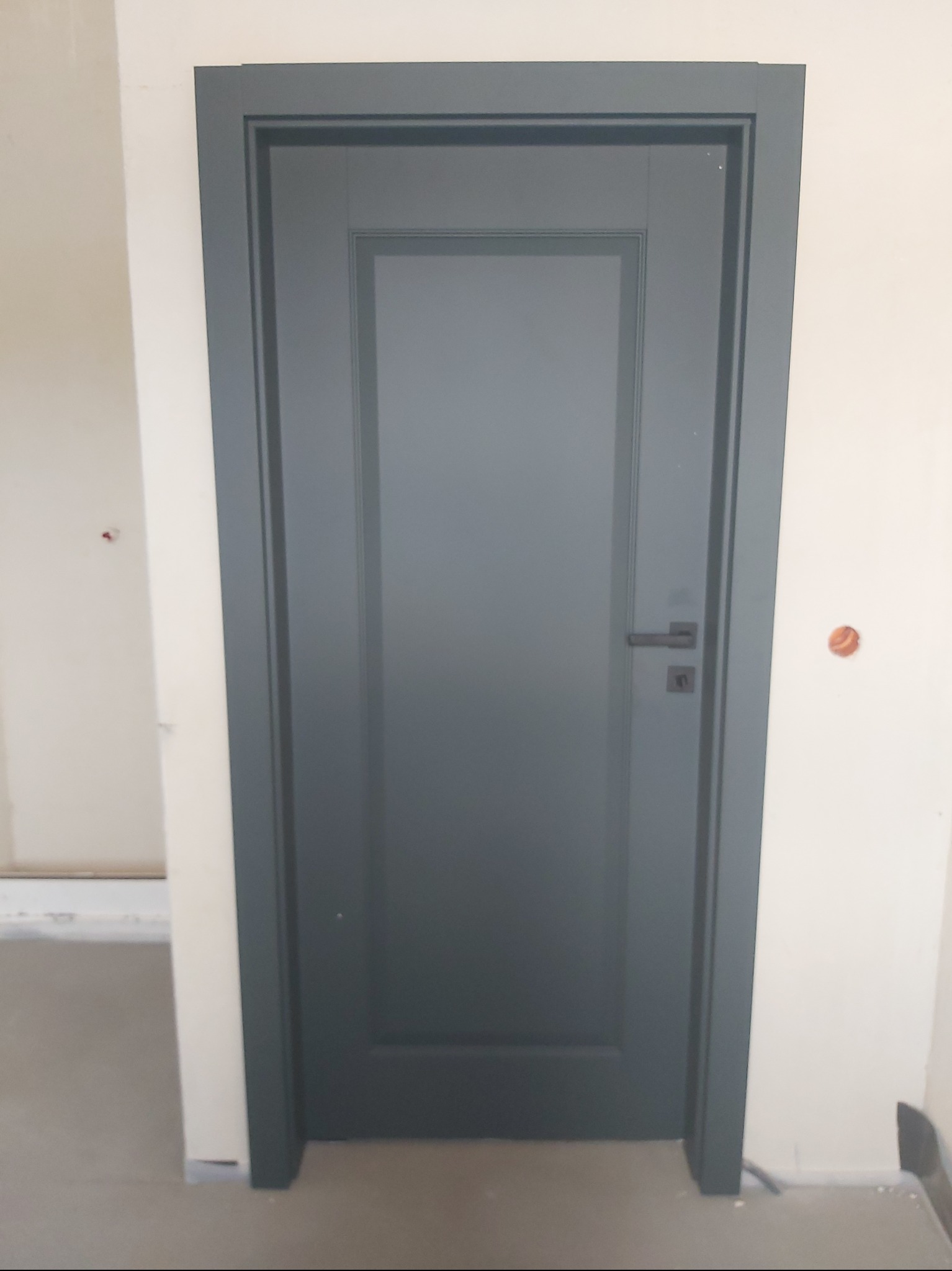 Zamknięte, grafitowe drzwi wewnętrzne z prostokątną ramą, czarną klamką i zamkiem, na tle jasnej ściany z widocznym otworem po gniazdku elektrycznym.