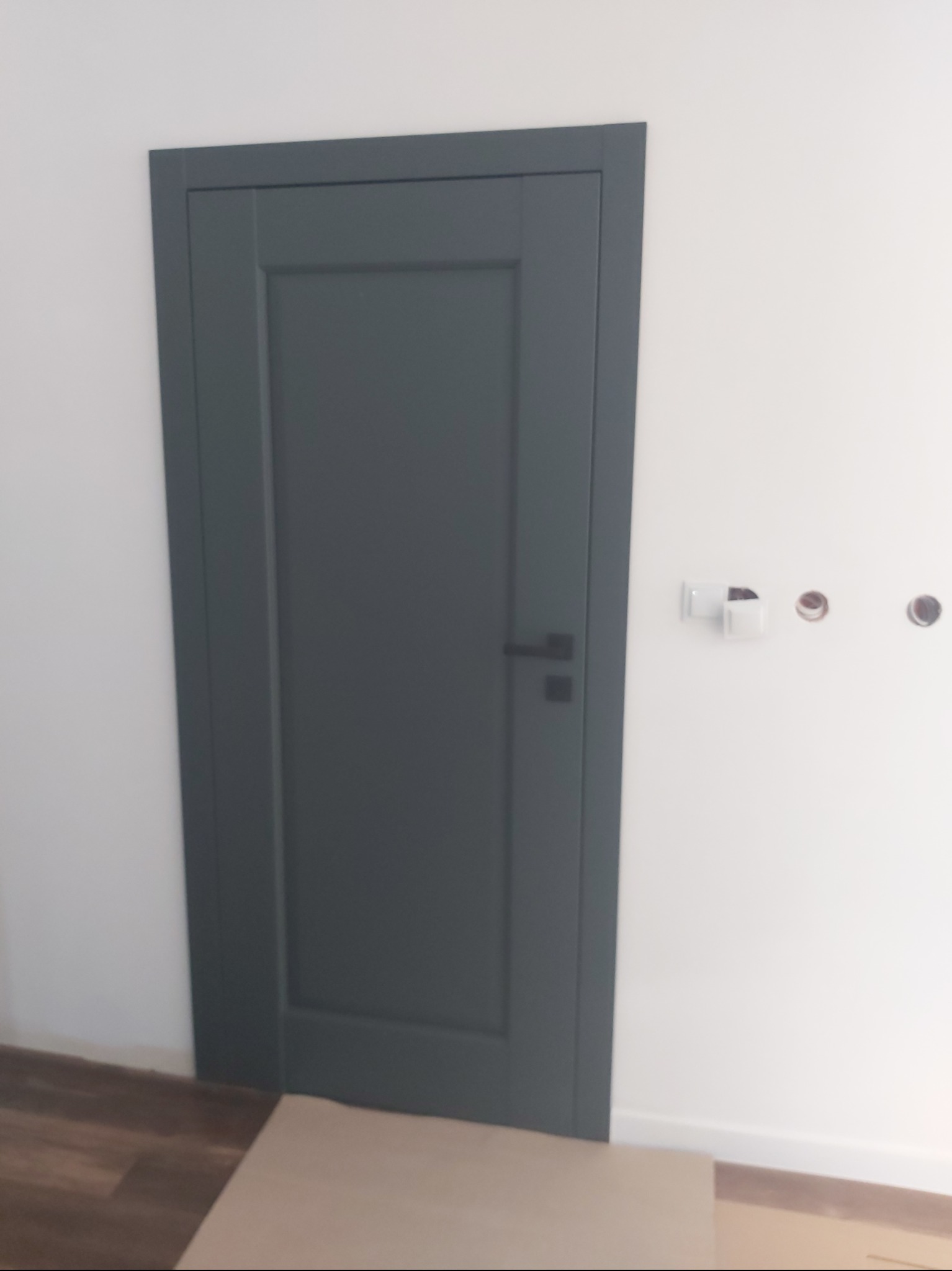 Zamknięte, grafitowe drzwi wewnętrzne z czarną klamką, osadzone w białej ścianie. Widoczne przygotowania do montażu osprzętu elektrycznego.