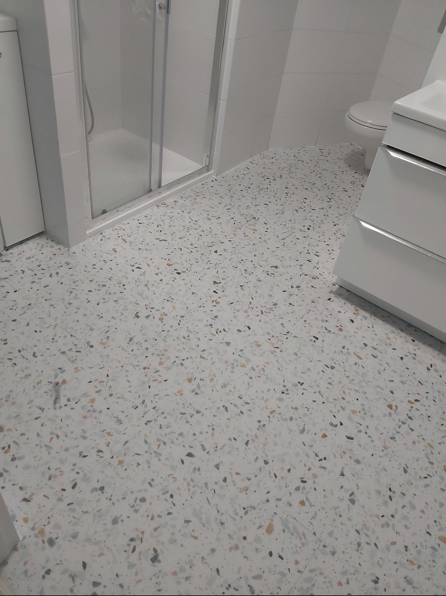 Jasna posadzka lastryko w łazience z kabiną prysznicową i umywalką. Nowoczesny wzór z drobnymi, kolorowymi kamyczkami. Minimalistyczny design.