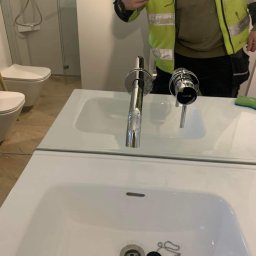 Marek Strzemiński - Nowa, chromowana bateria umywalkowa marki Kludi zamontowana w białej umywalce. Widoczne odbicie hydraulika w kamizelce podczas robienia zdjęcia telefonem.