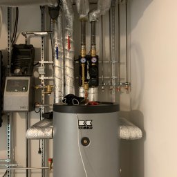 Marek Strzemiński - Szary zasobnik ciepłej wody REMKO z widocznymi instalacjami, rurami w izolacji i manometrami. Na zasobniku leżą rękawice robocze i narzędzia.