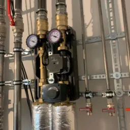 Węzeł cieplny z dwoma manometrami, pompą obiegową Grundfos i srebrną izolacją termiczną rur. Instalacja przymocowana do ściany za pomocą metalowych uchwytów.