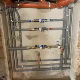 Szare rury instalacji hydraulicznej z zaworami i manometrami, zamocowane do ściany z widocznymi śladami wilgoci. U góry rury kanalizacyjne z pomarańczowego tworzywa.